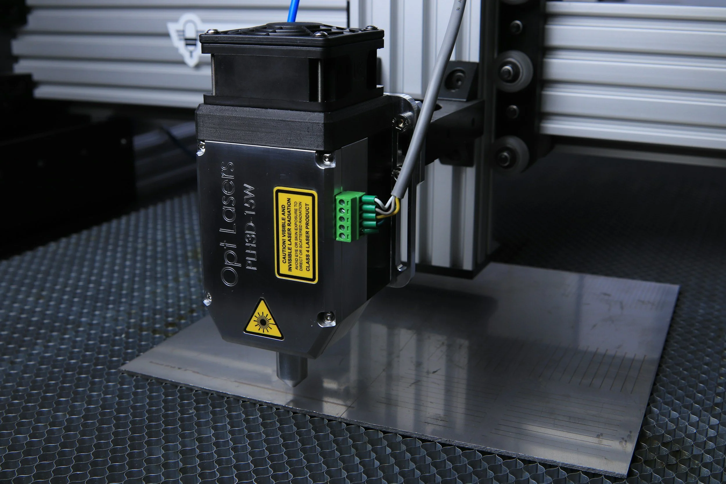 Close-up of a laser engraving or cutting machine with black and metallic components, yellow caution labels, and wires connected, operating on a flat surface with a textured pattern.