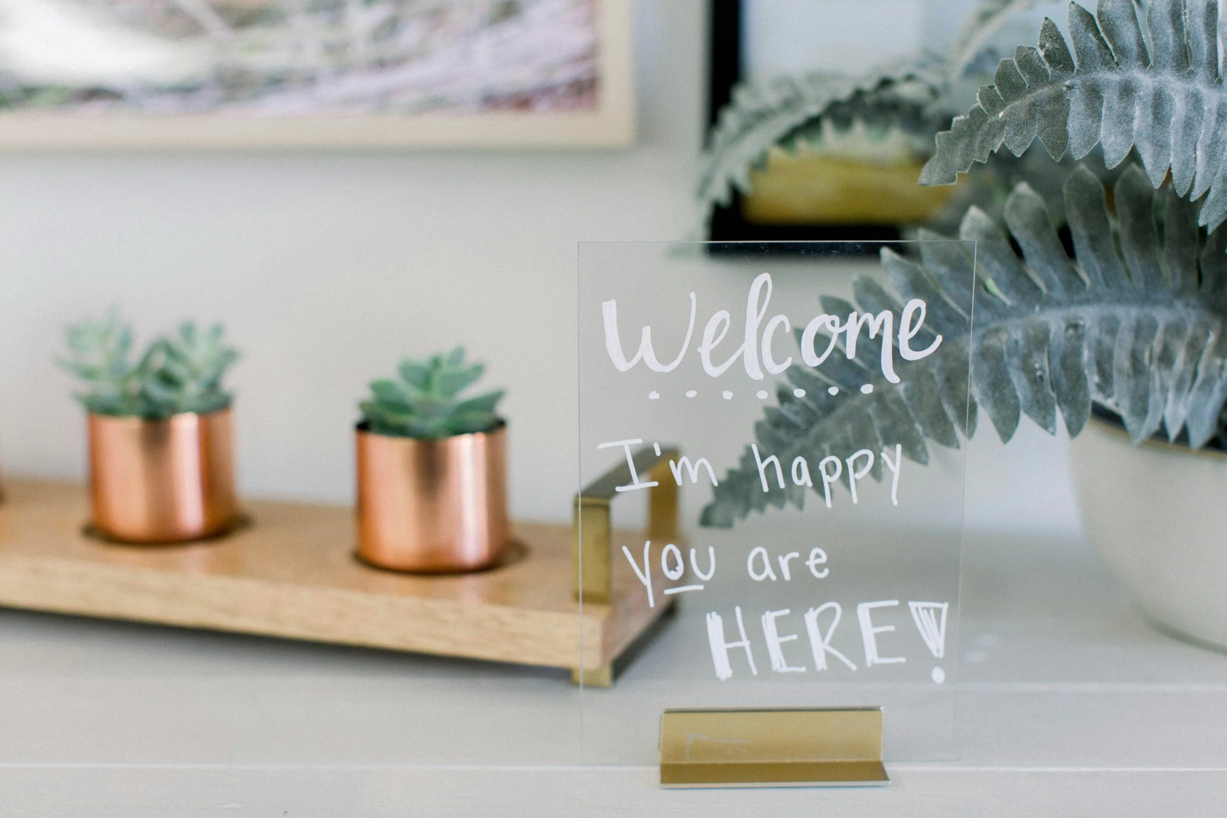 Close-up of a decorative glass sign with handwritten style text that reads 'Welcome, I'm happy you are here,' positioned in front of a plant and a shelf with small potted succulents in copper containers.