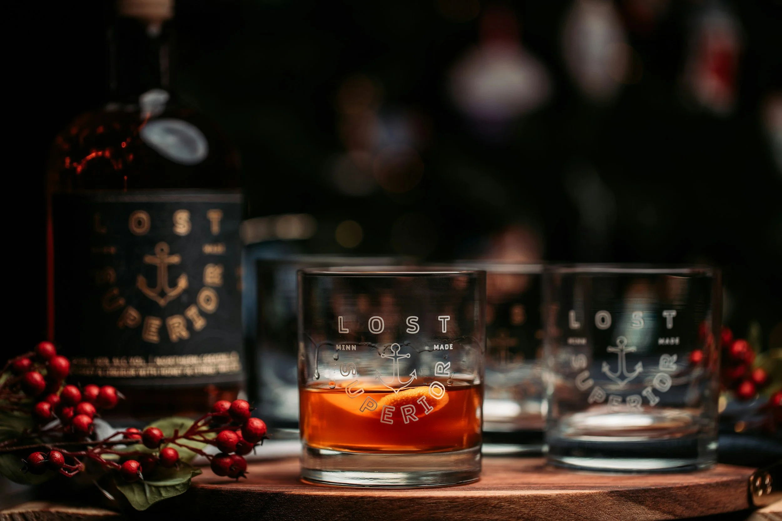 Close-up of a glass with a drink, with a background of dark bottles. There is holiday decor with red berries and holly leaves in the foreground.