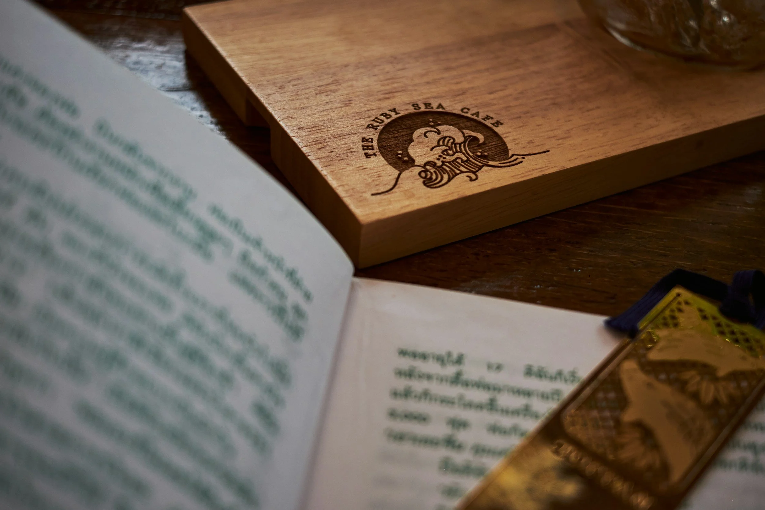 A wooden coaster with a logo reading 'The Ruby Sea Cafe' and an illustration of a wave. An open book with blurry text is in the foreground, and part of a glass container is visible at the top right corner.