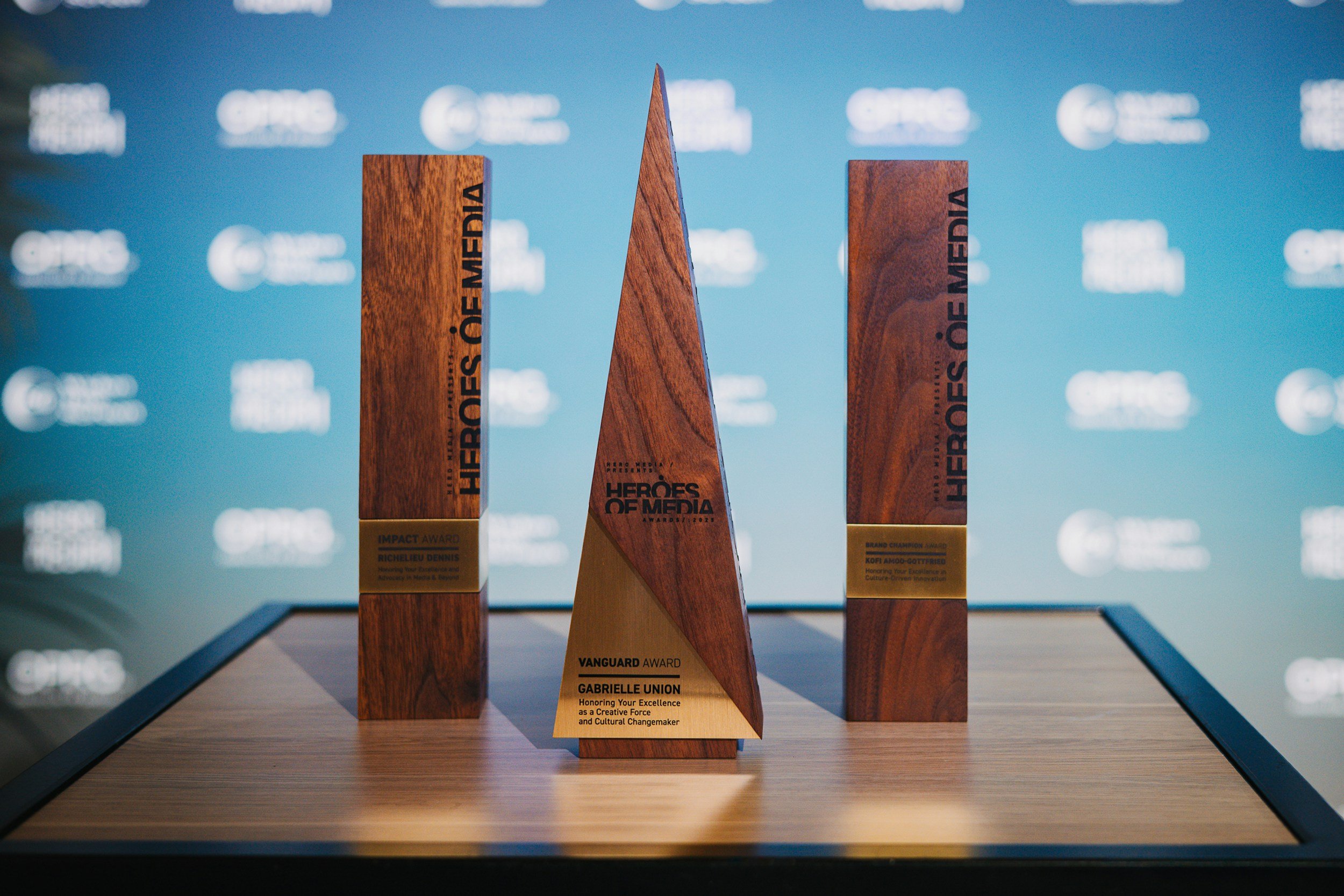 Three wooden awards on a display table, with visible plaques indicating the names of recipients and awards, set against a blue background with blurred event branding.