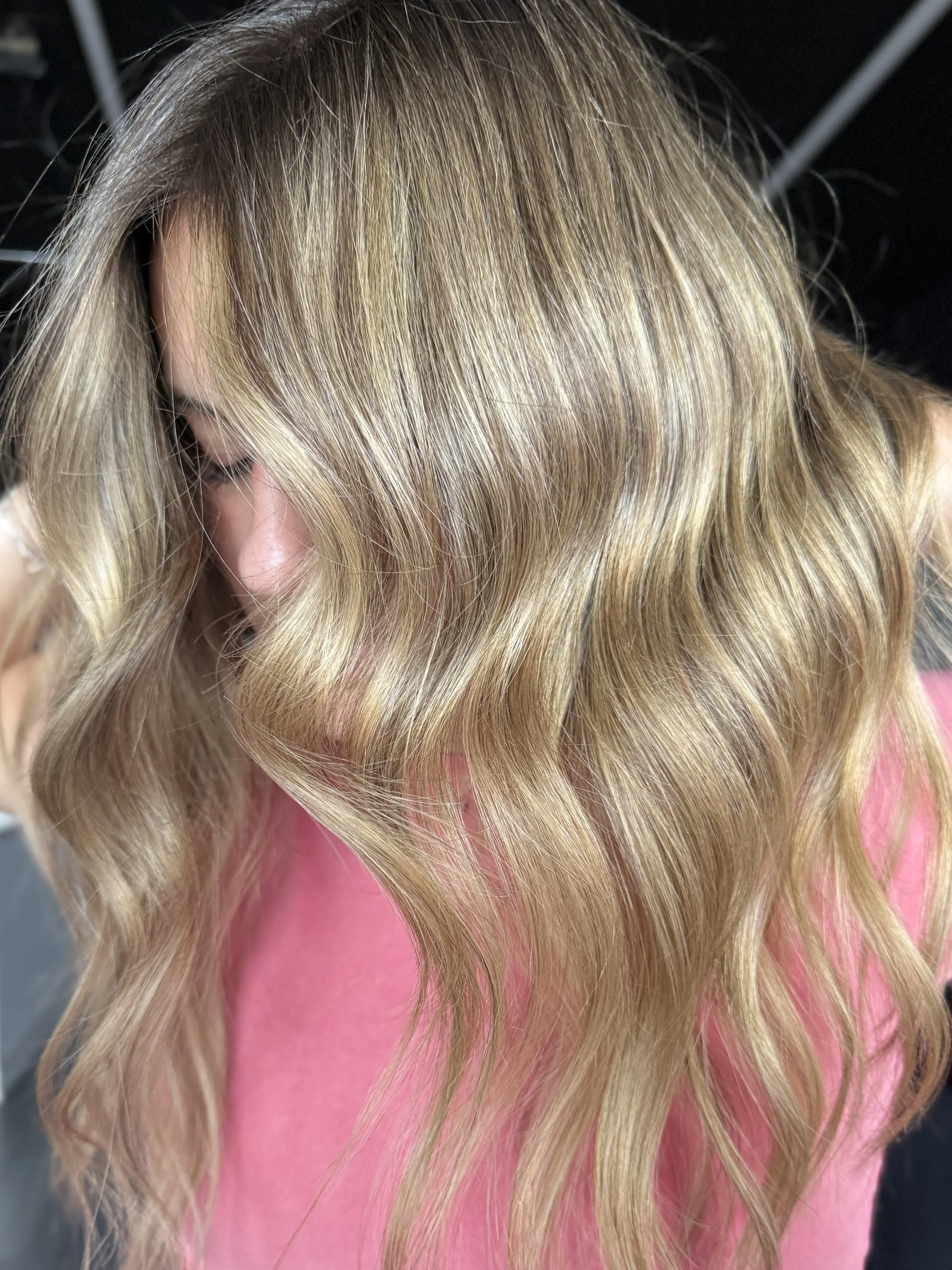 Close-up of blonde hair styled in loose waves, wearing a pink top.