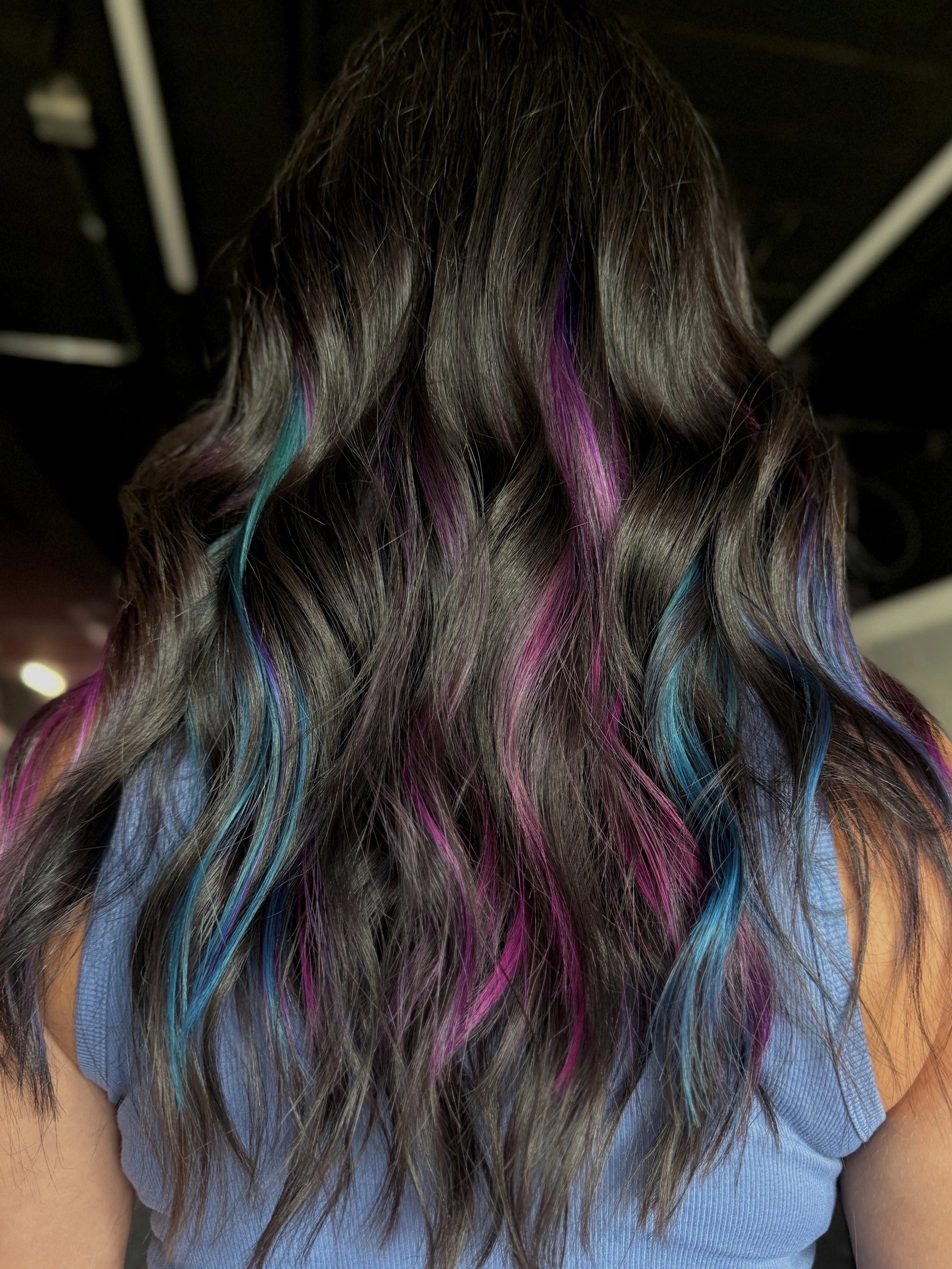 Back view of a person with long, wavy hair dyed with streaks of purple, pink, and blue, wearing a blue shirt.