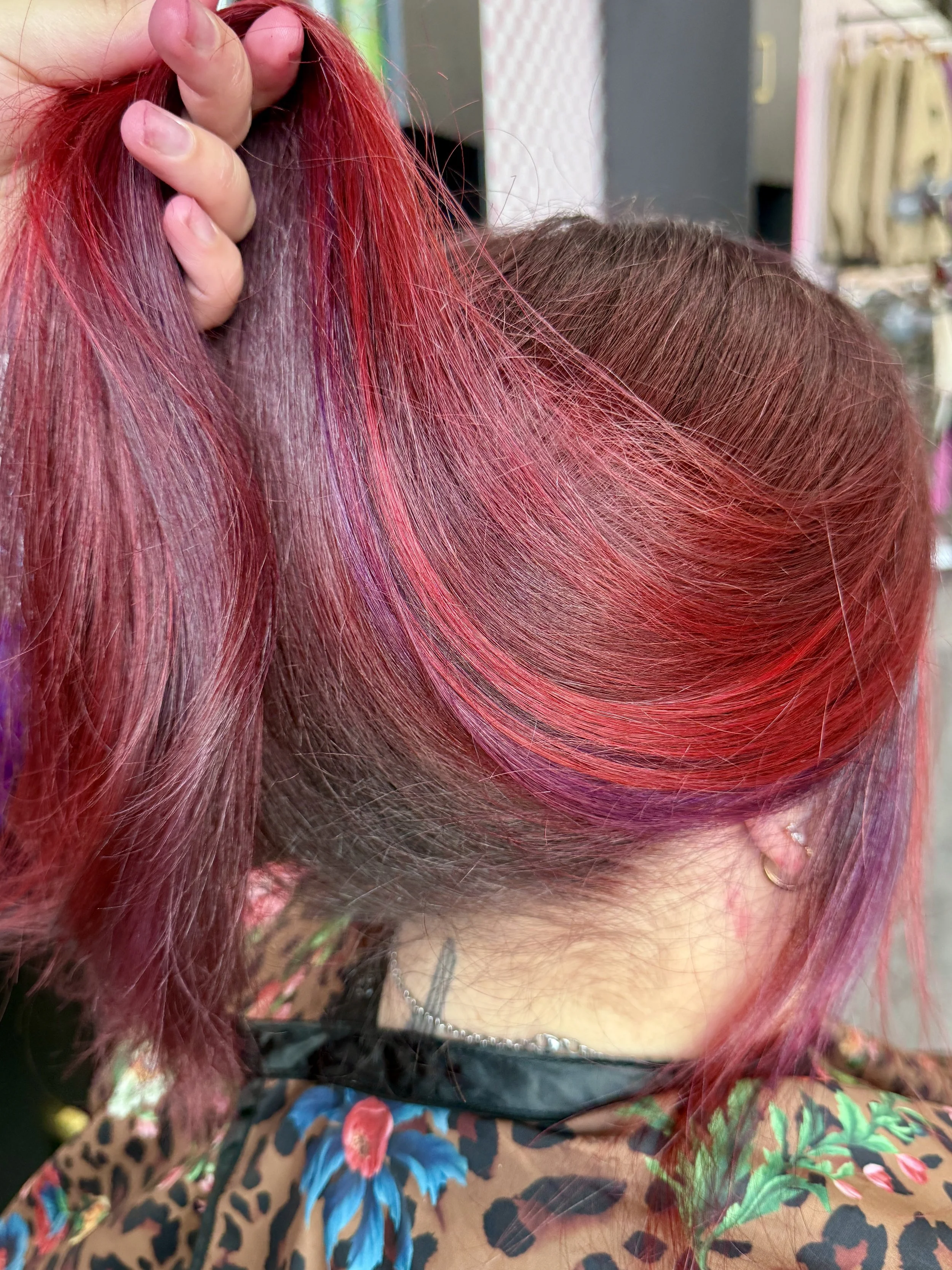 Close-up of a person's hair with bright red and purple colors, styled with a hand holding a section of it, wearing floral and animal print clothing.