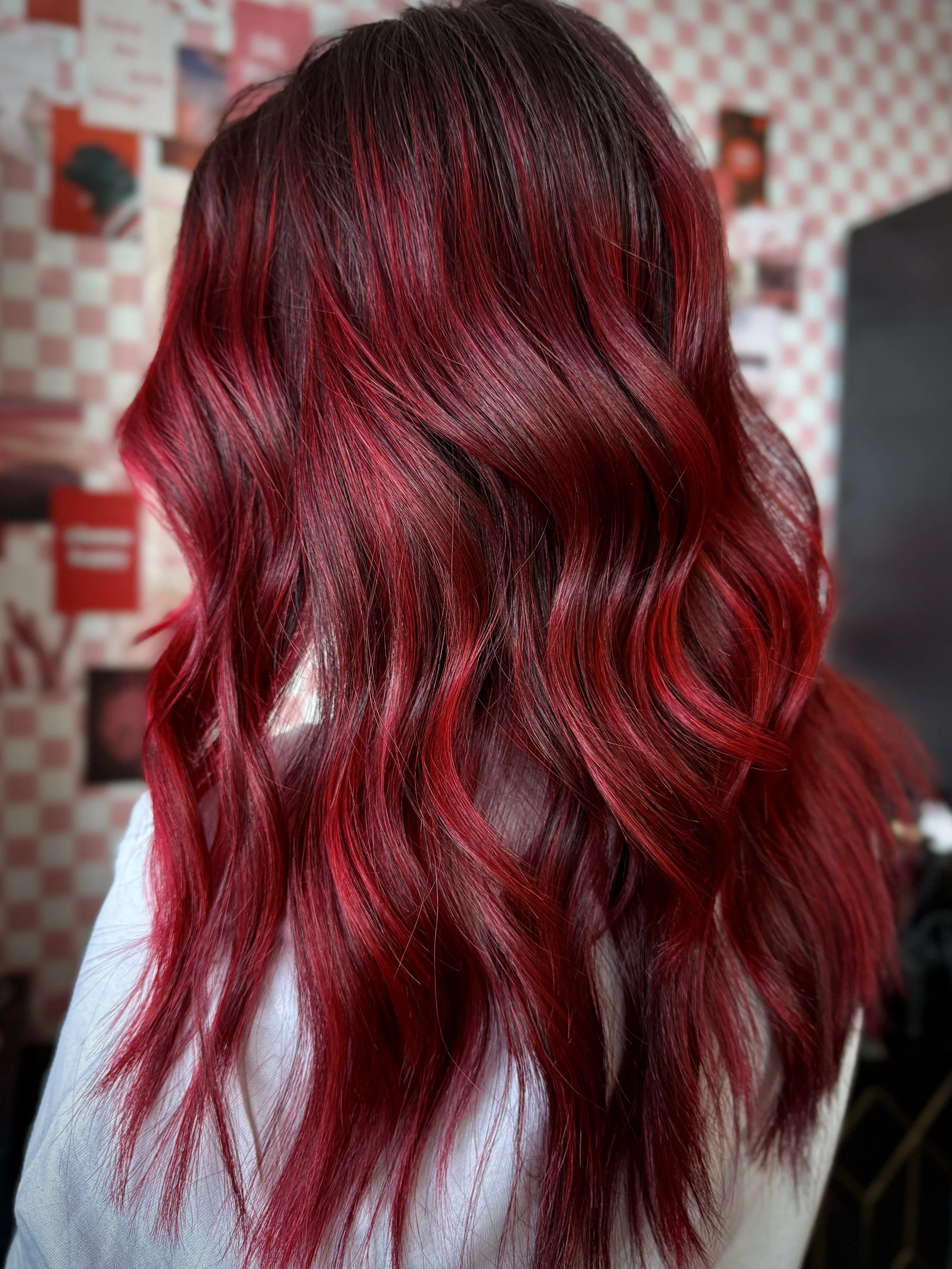 Person with long, wavy red hair standing in a salon with a red and white checkered wall in the background.