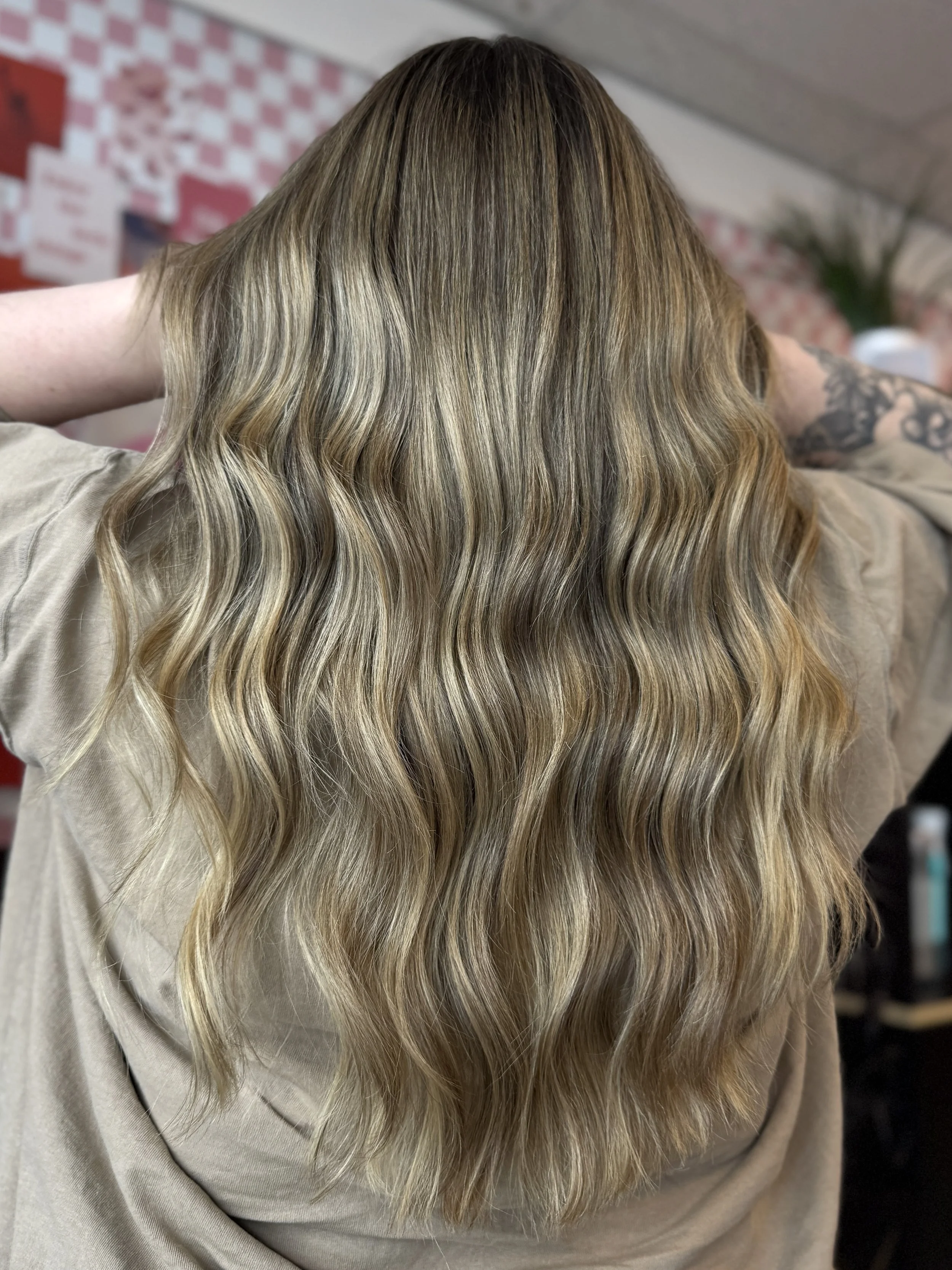 Close-up of a person with long, wavy, blonde hair, wearing a beige top, with a blurred background featuring a wall and some greenery.