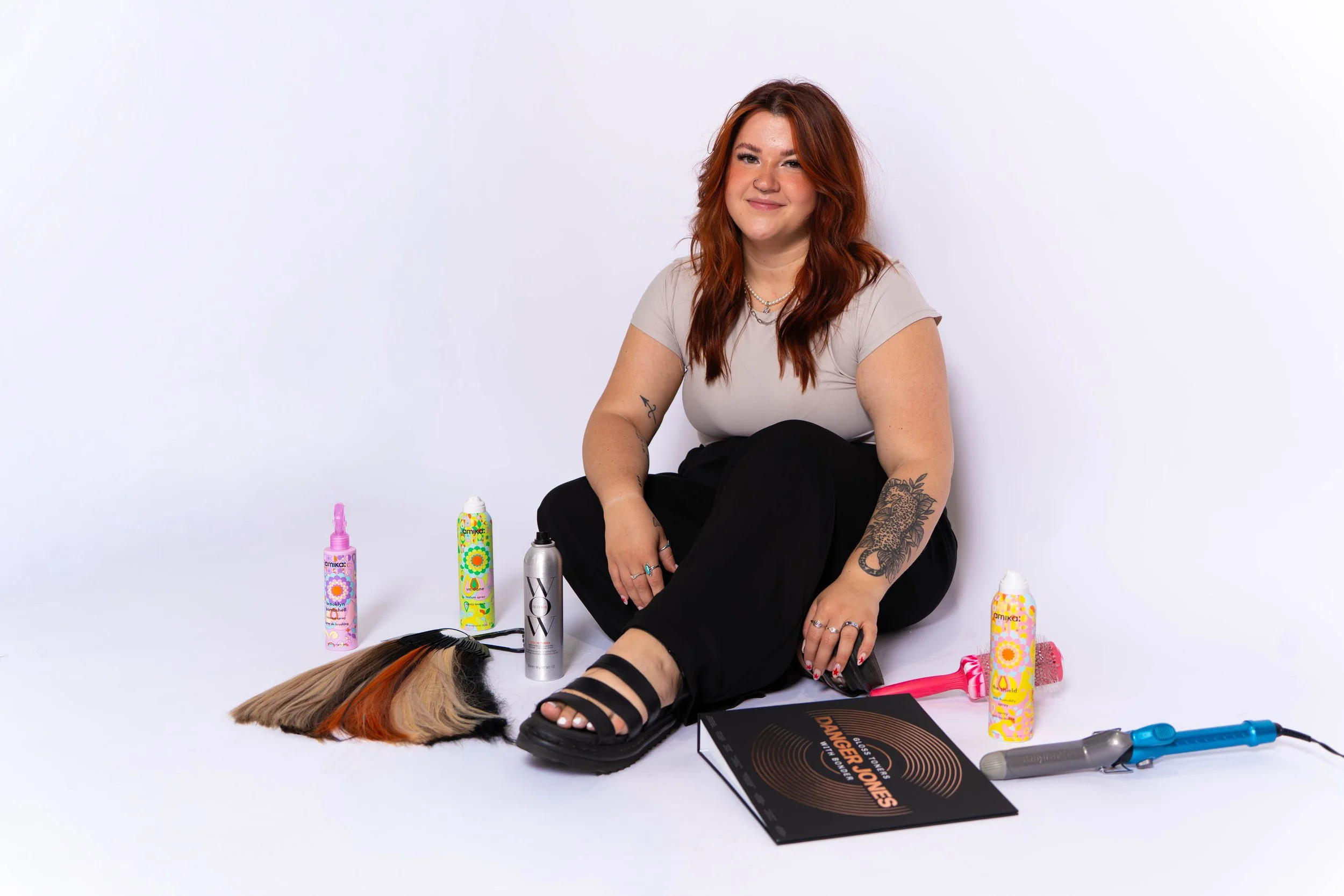 A woman with red hair sitting on the floor surrounded by hair styling tools and products. She is smiling and looking at the camera, with a colorful wig in front of her.