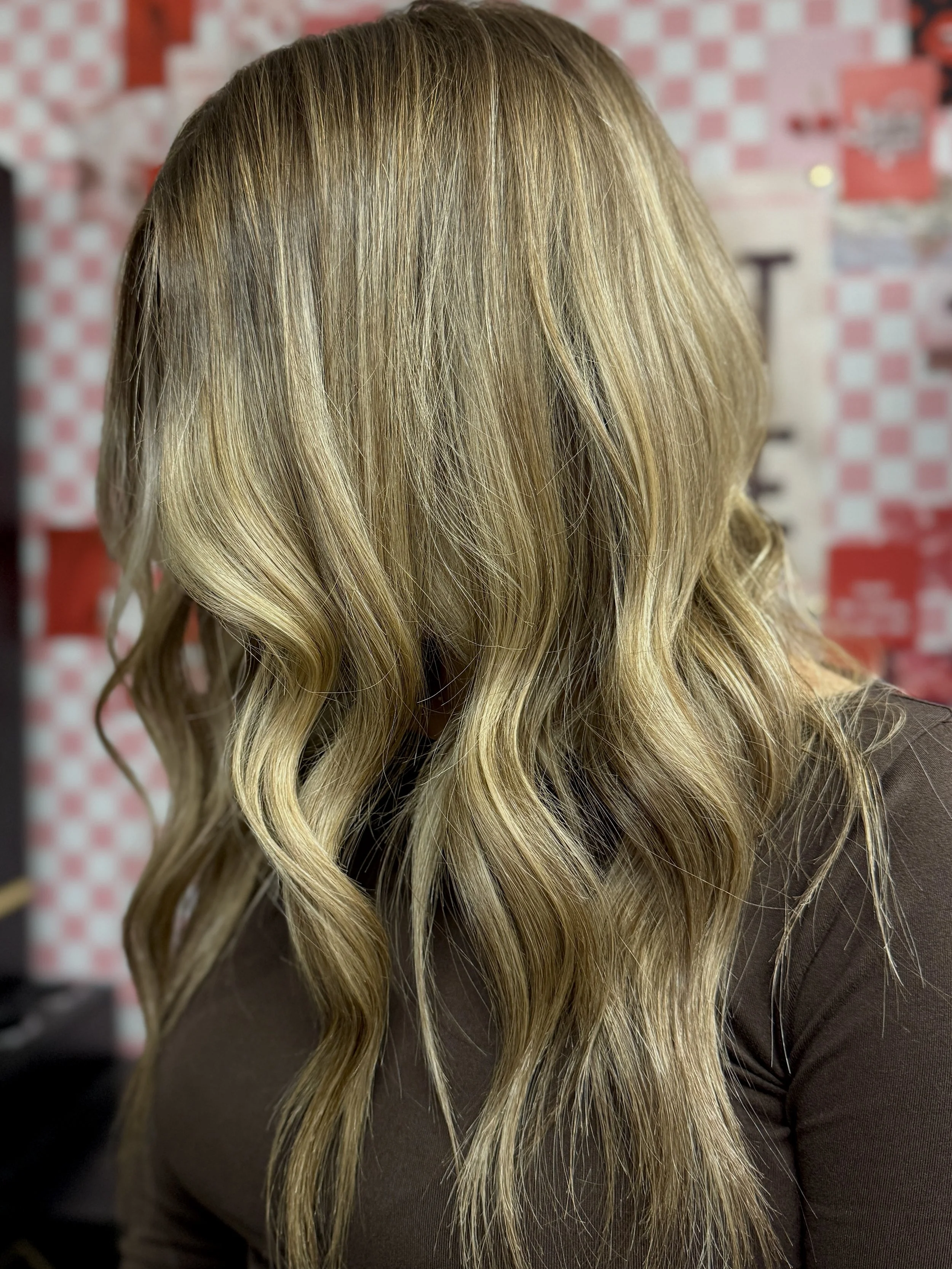 A close-up of long, wavy blonde hair with subtle highlights, wearing a dark top, with a red and white checkered patterned background.