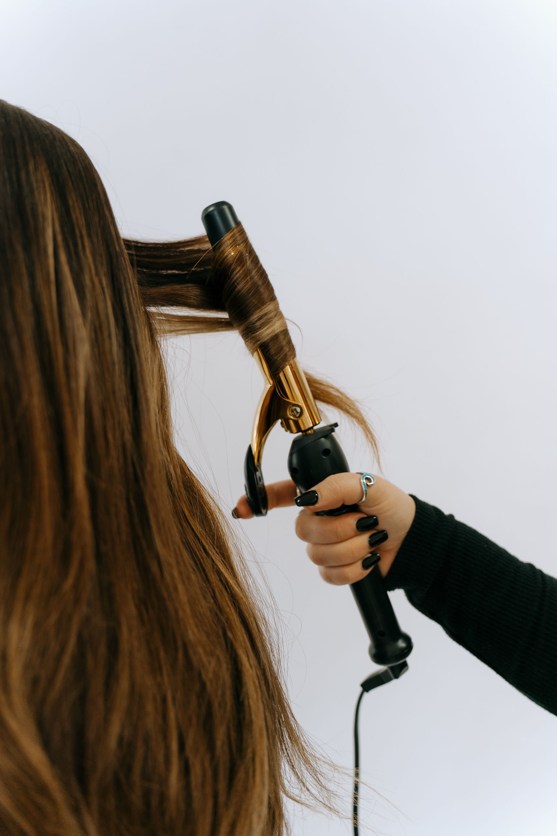 A person with long, red-brown hair is curling their hair with a curling iron. The hand holding the curling iron is wearing black nail polish and a silver ring.