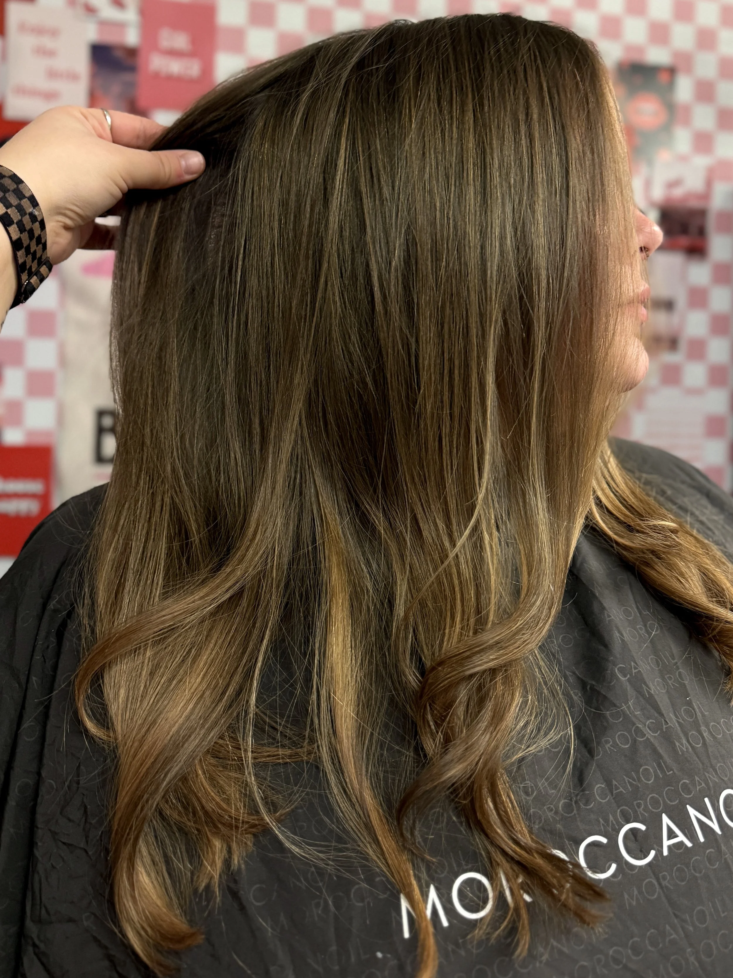 A woman with long, wavy, brown hair is seated in a salon chair, with a hand touching her hair. The background features a red and white checkered pattern and various salon promotional posters.