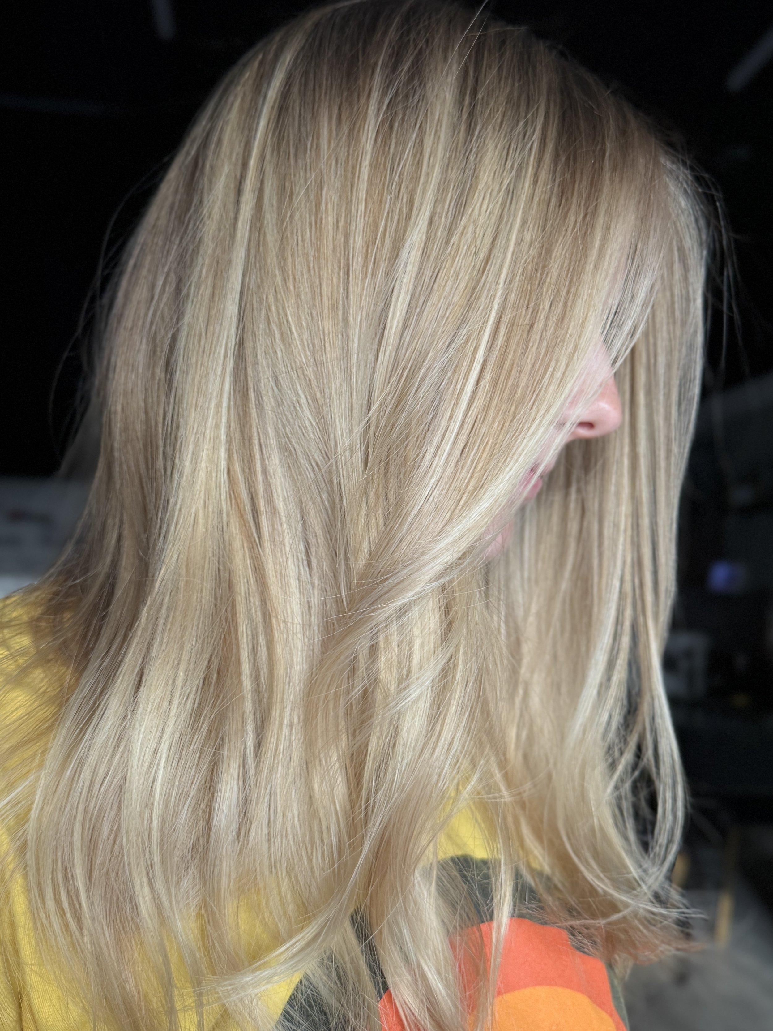 Close-up of a woman with long, straight, blonde hair wearing a yellow and orange patterned top. The background is dark and out of focus.