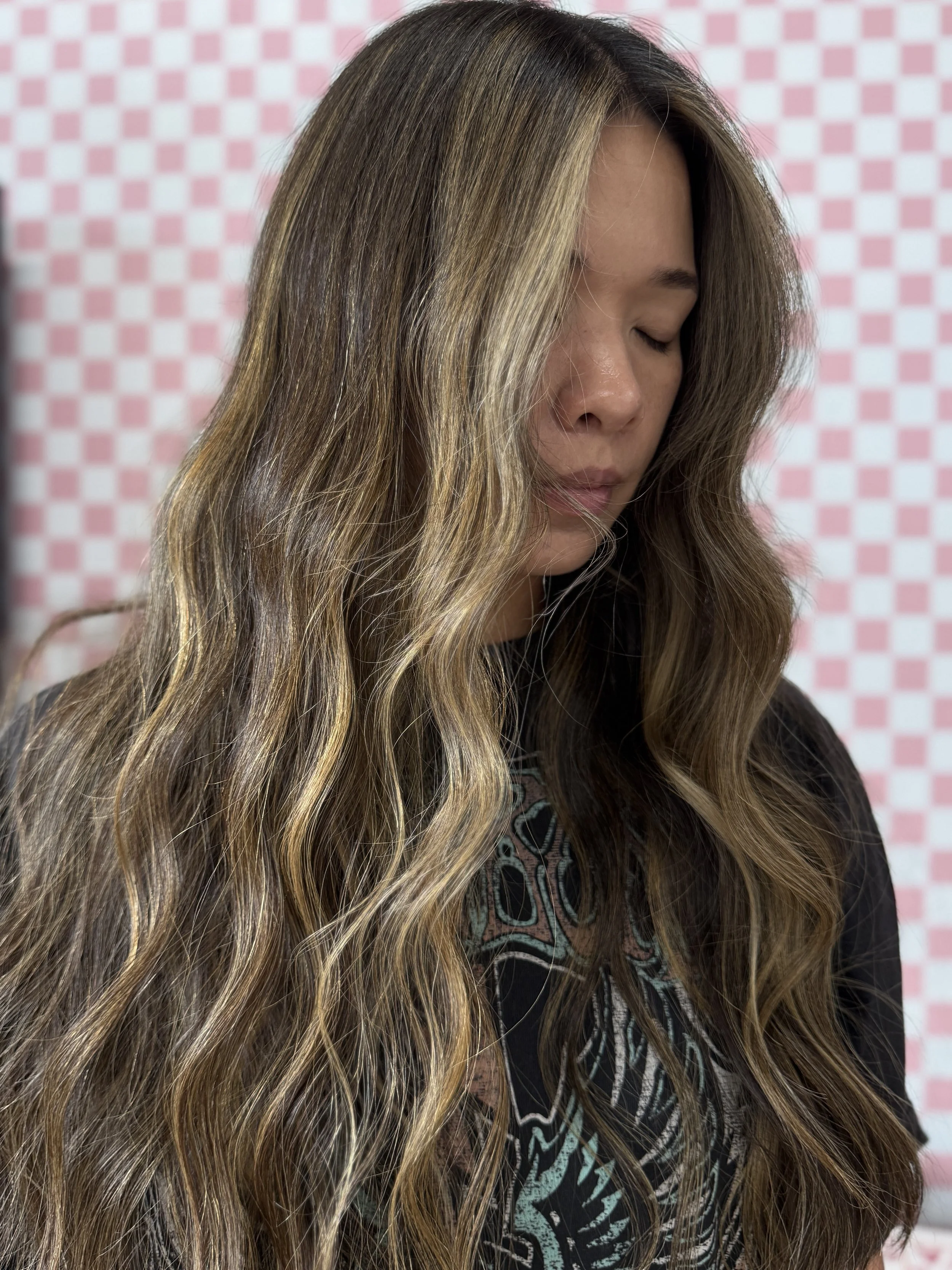 A woman with long, wavy hair with blonde highlights, facing downward with eyes closed, against a pink and white checkered background.