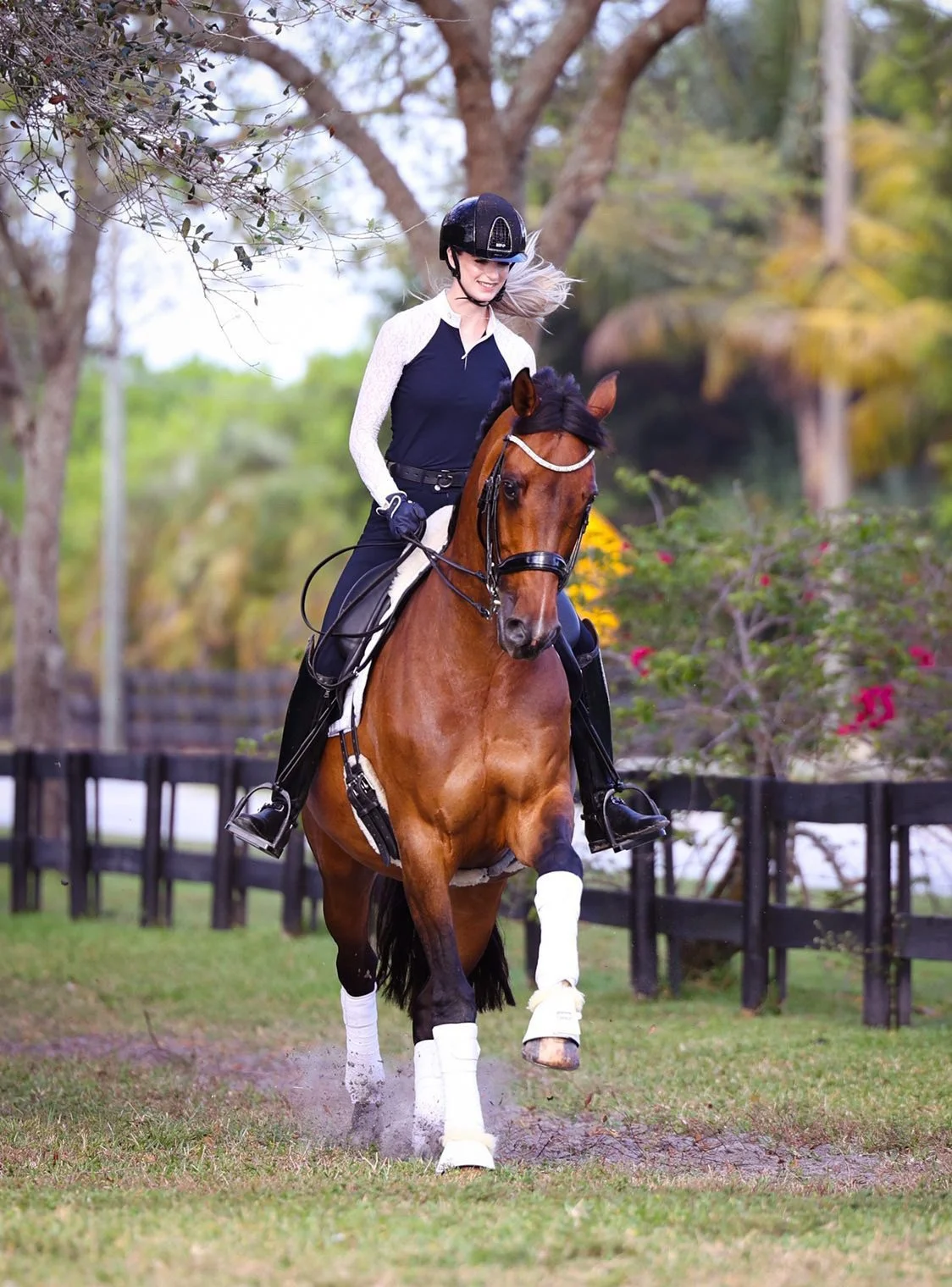 A woman riding a brown horse on a grassy field, wearing a black riding helmet, a black and white riding jacket, black pants, and gloves, with trees and a black fence in the background.
