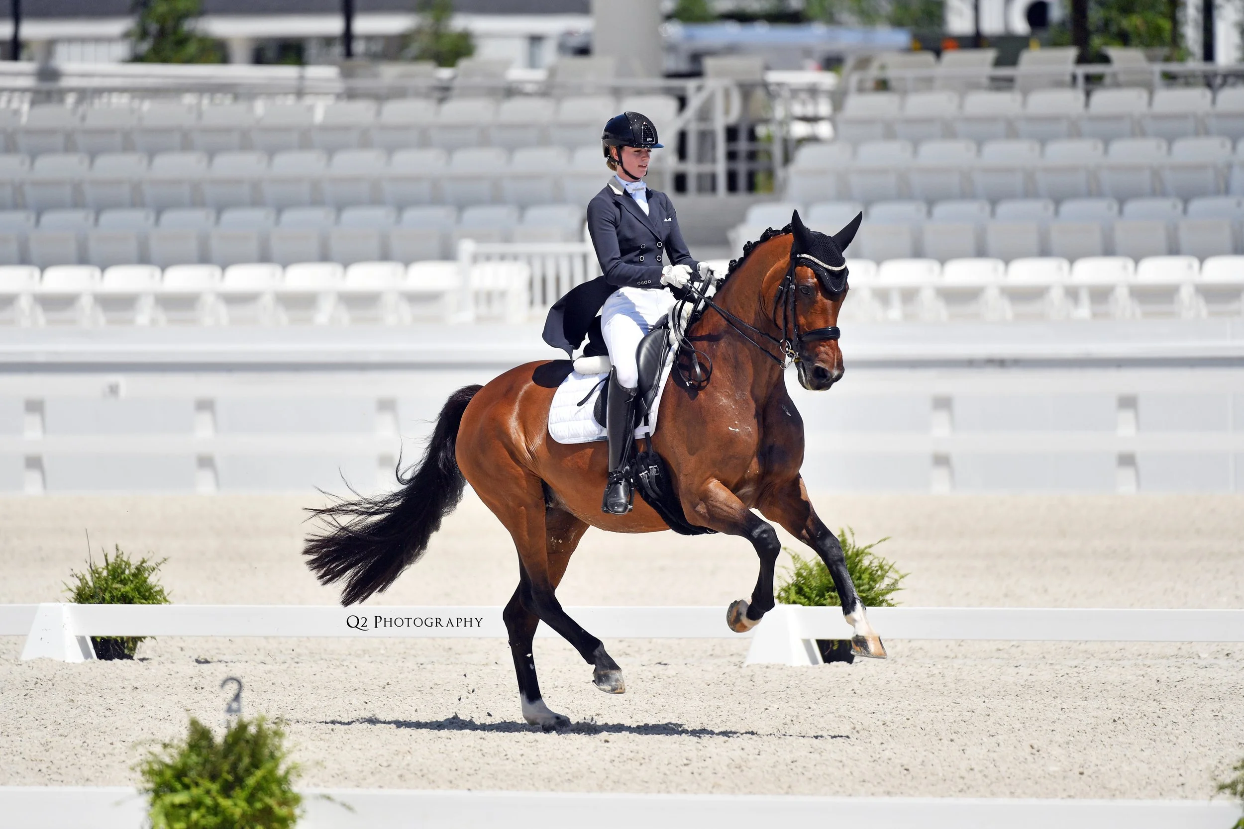 A female equestrian rider in formal riding attire, including a helmet, jacket, and white breeches, riding a brown horse with a black mane in a dressage arena with white barriers and empty bleachers in the background.