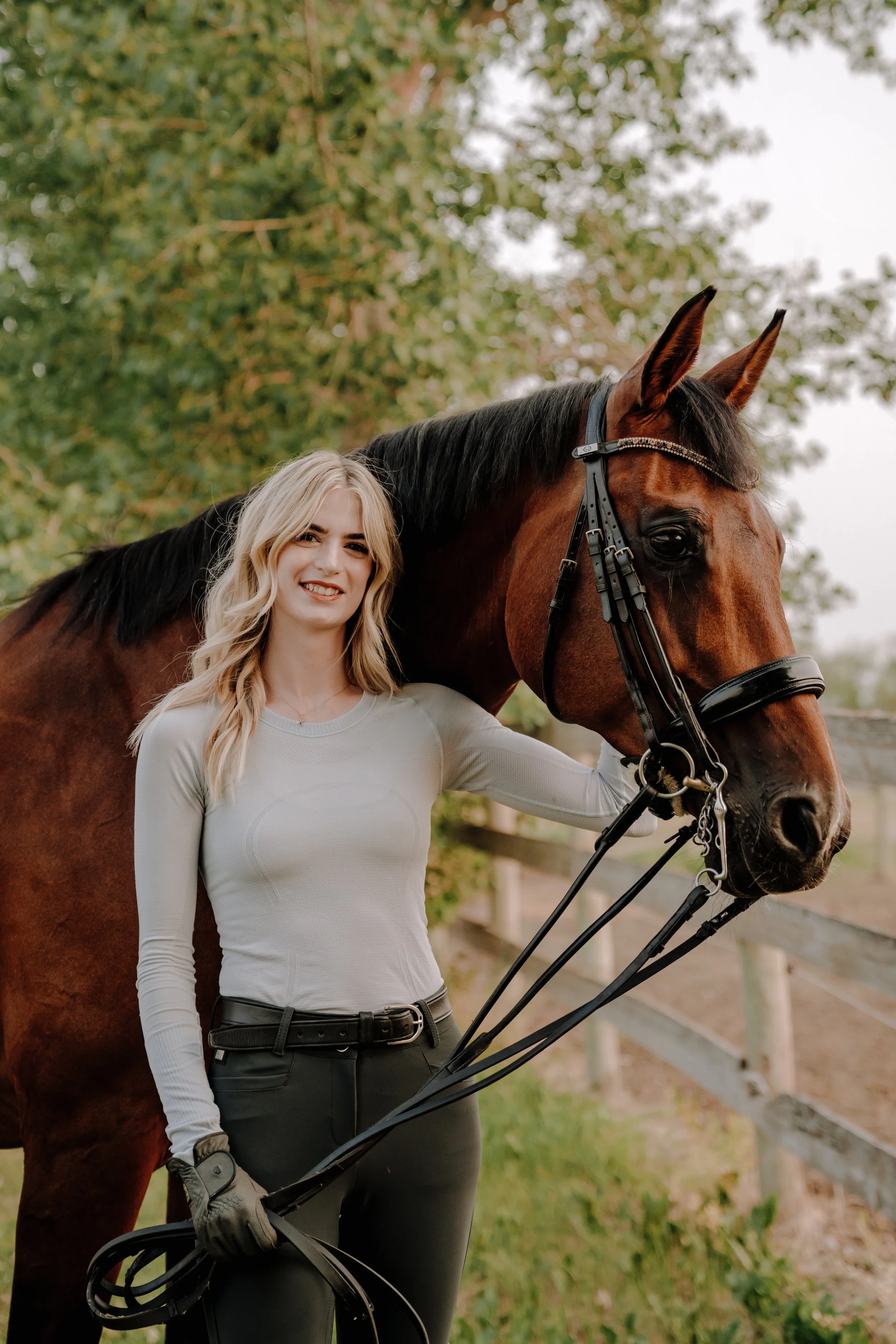A young woman with long blonde hair smiling and standing next to a brown horse with a black mane outdoors, with trees and a white fence in the background.