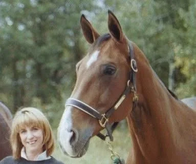 A girl with blonde hair smiling next to a brown horse with a white blaze on its face in an outdoor setting.