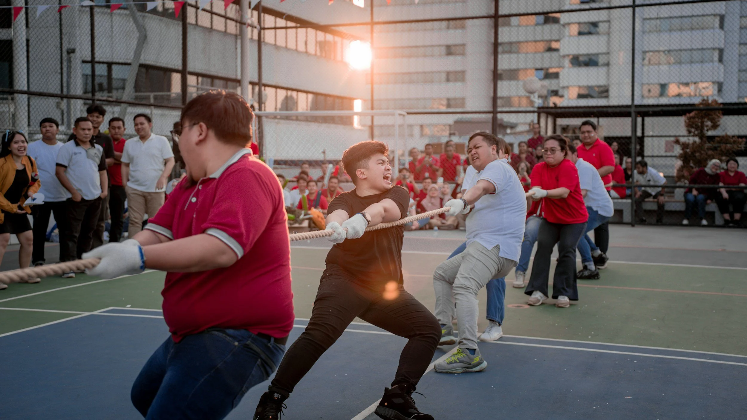 Teams compete in a tug-of-war on an outdoor court as spectators watch in the sunlight during an adult Messy Play Party.