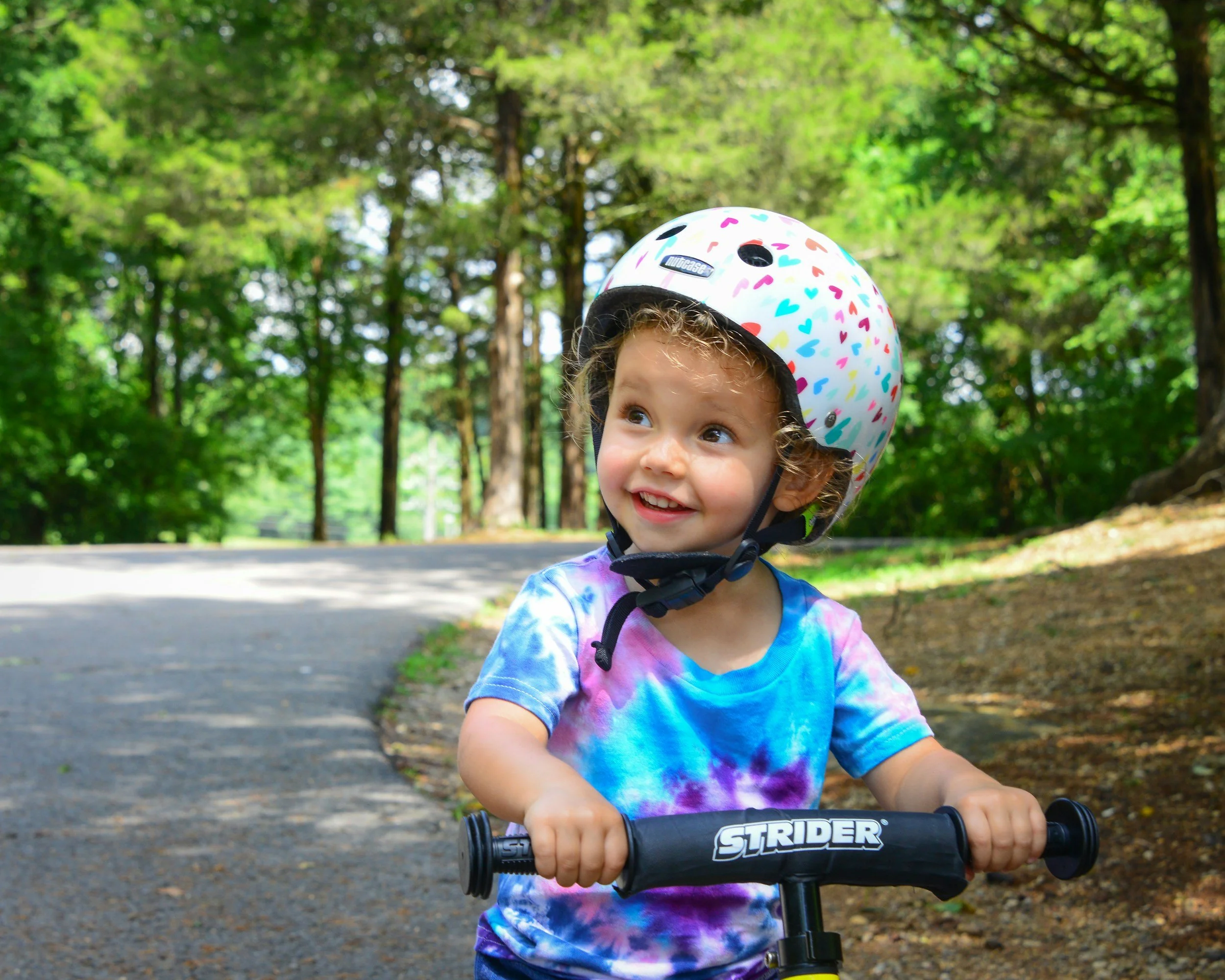 Smiling child wearing a colorful helmet and riding a balance bike on a wooded path, heading to a Messy Play tie-dye party