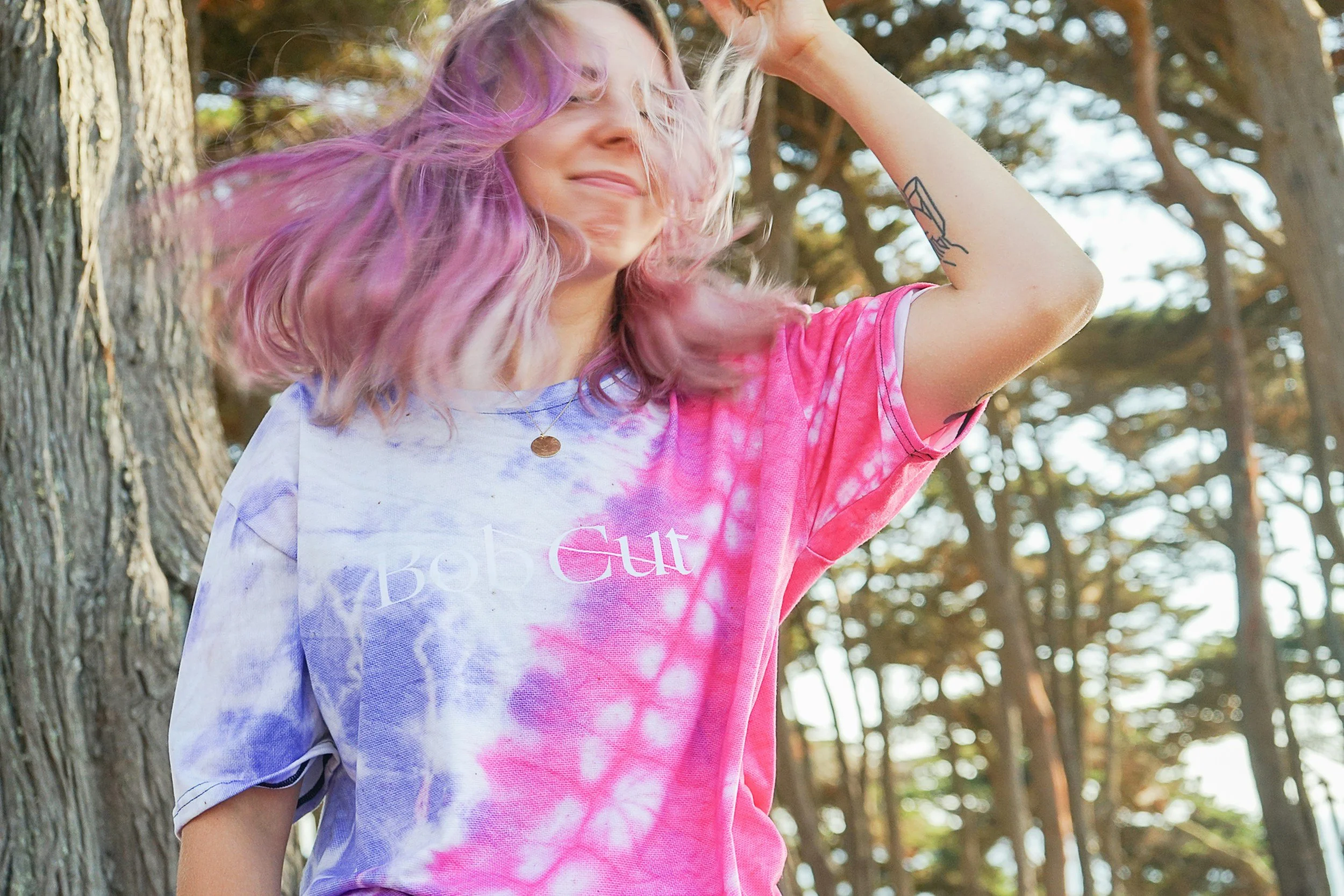 Adult with flowing pink and purple hair posing beside a tree in a sunlit forest during a Messy Play Tie-Dye Party