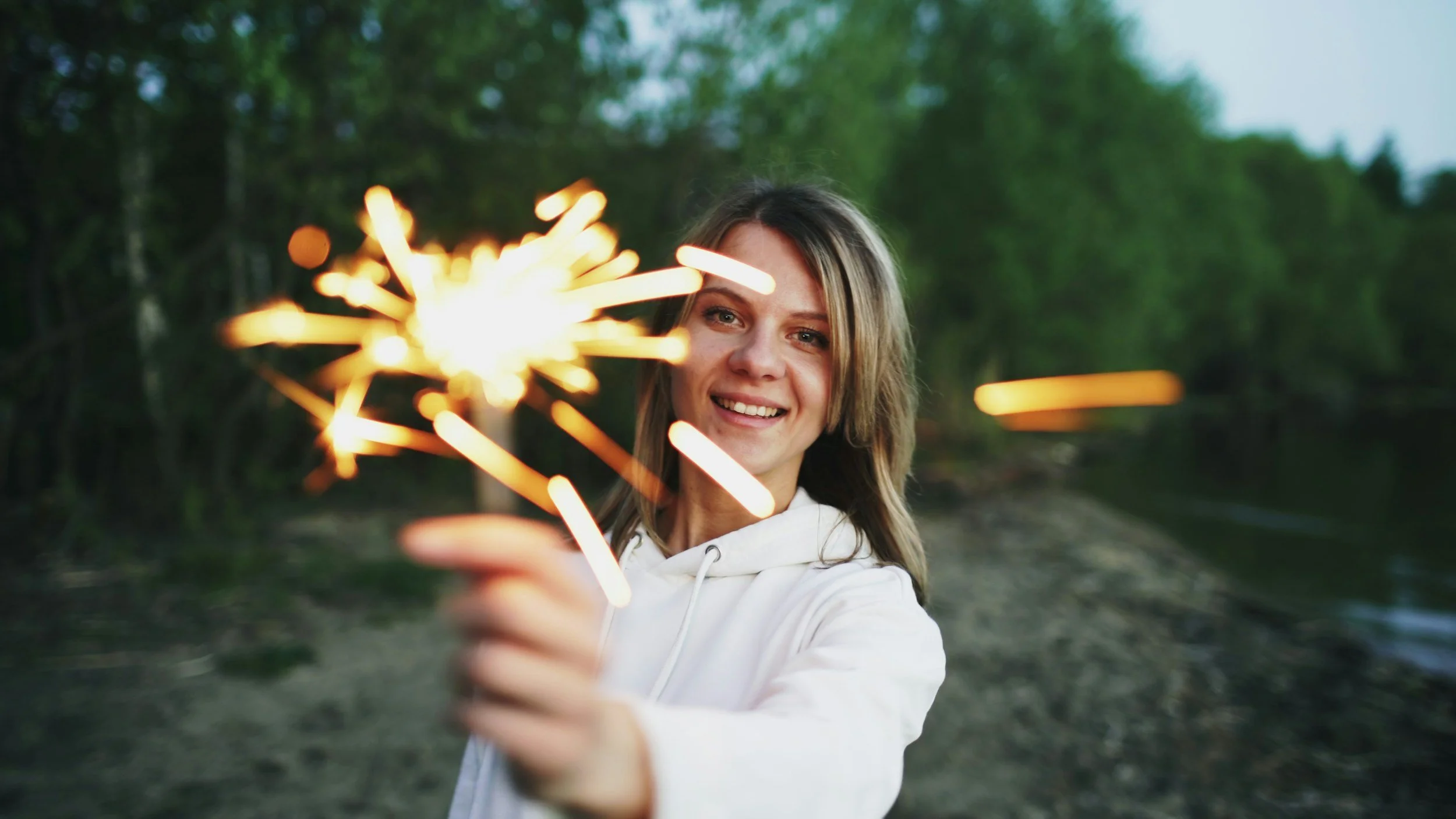 Smiling woman in a white hoodie holding a glowing sparkler outdoors in a forest