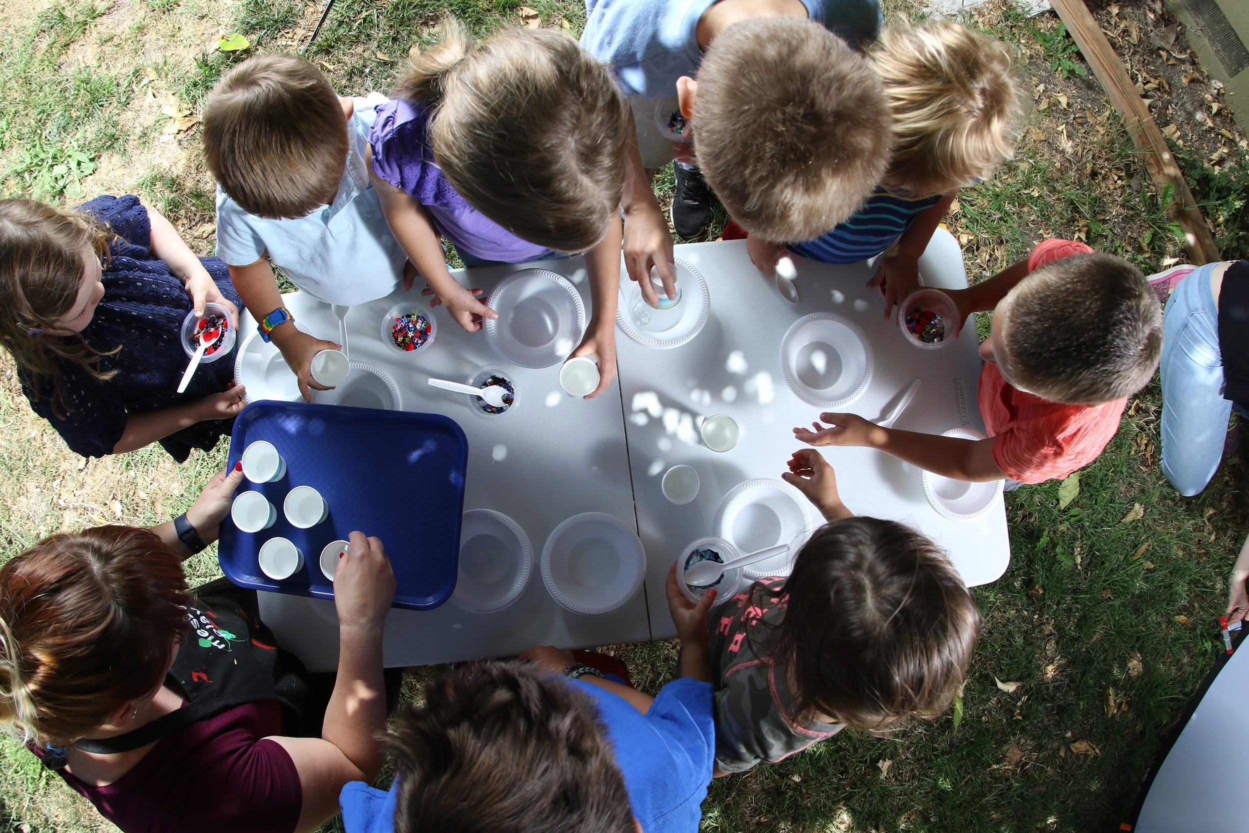 Overhead view of children making crafts with colorful beads, bowls, spoons, and cups at an outdoor table.