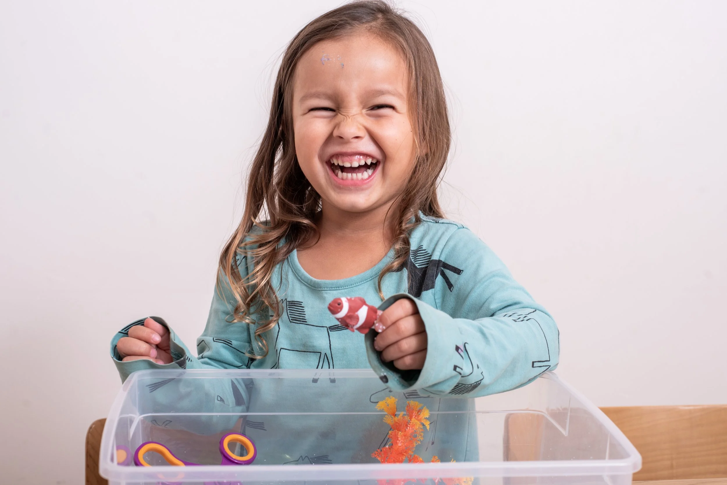 Smiling young girl playing with a red toy fish in a sensory bin.