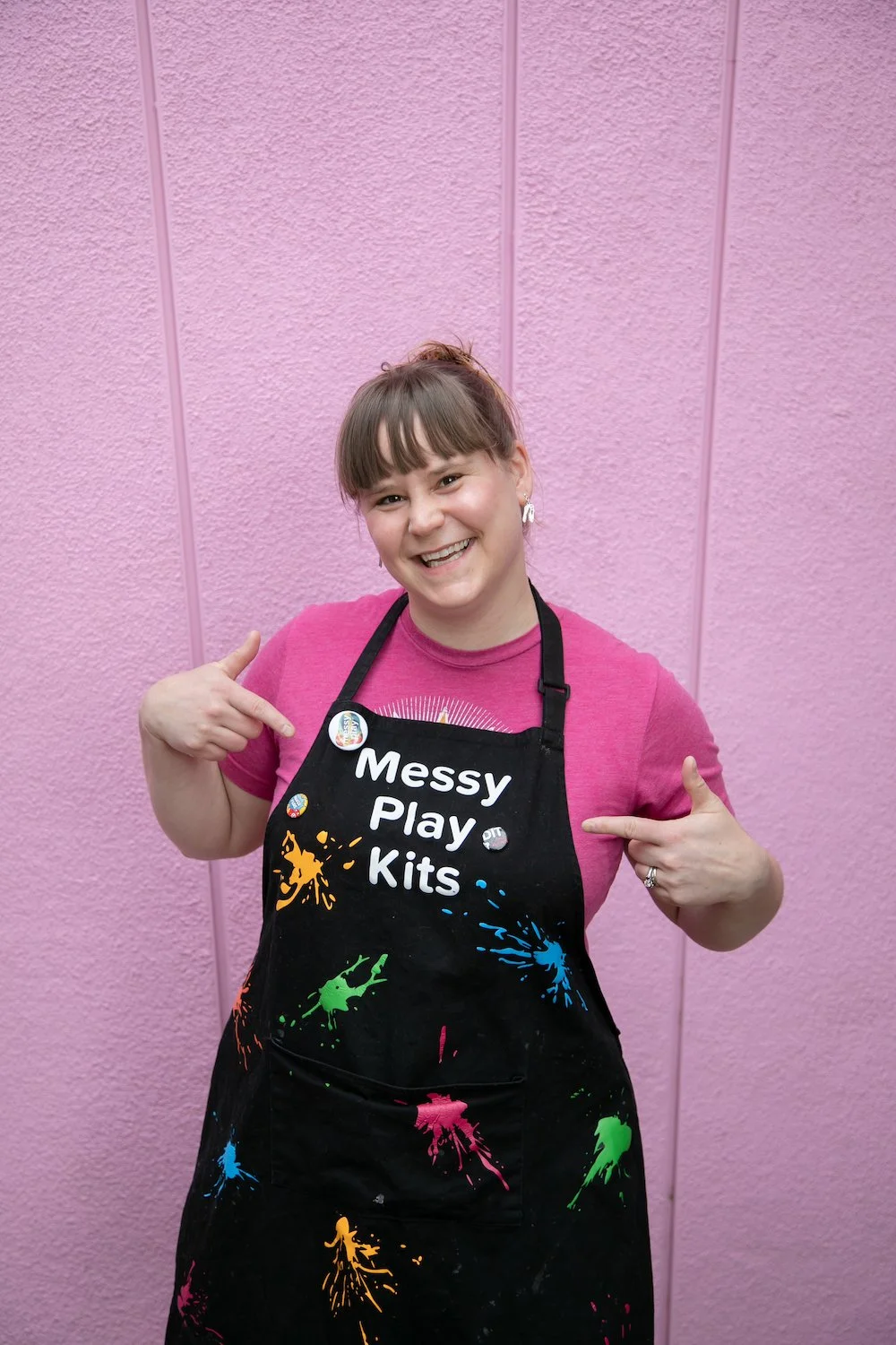 Robin Imholte pointing to a colorful black “Messy Play Kits” apron against a pink wall