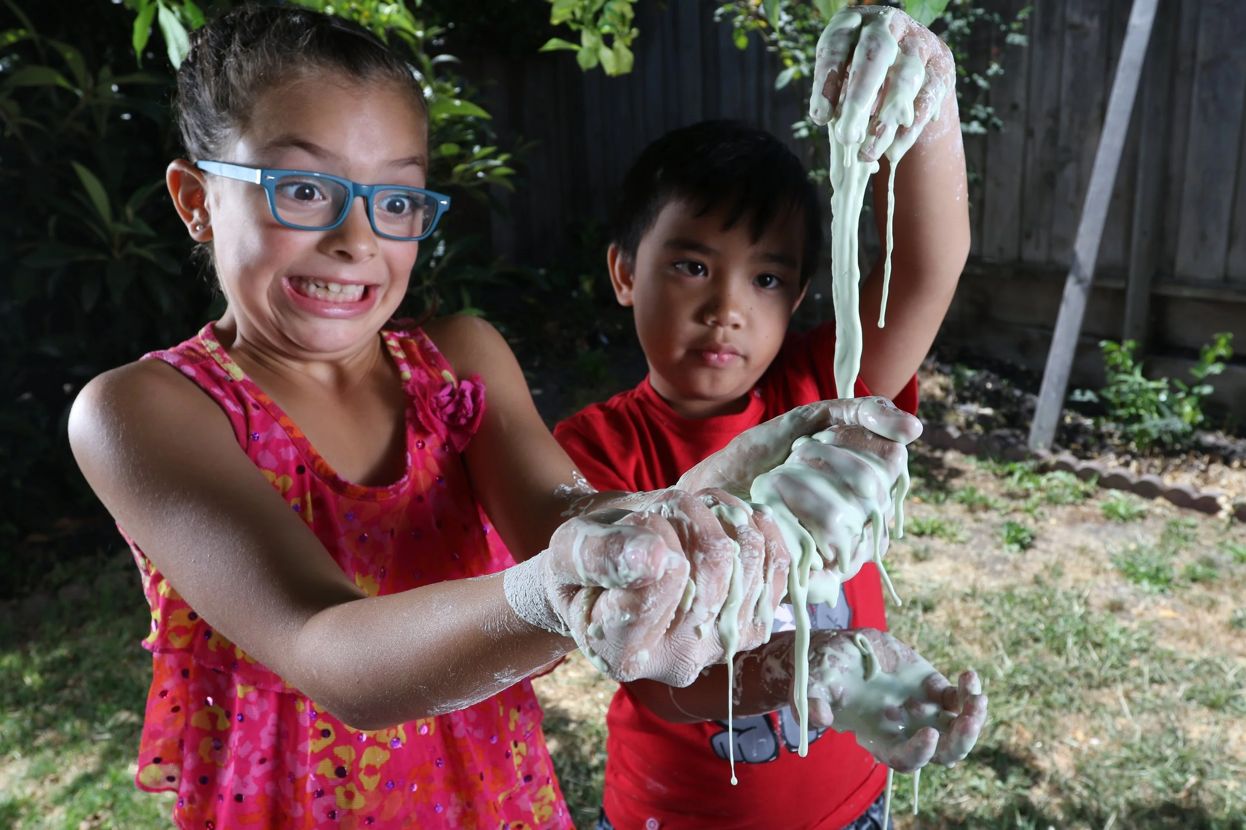Two children holding stretchy green slime outdoors, with slime covering their hands and dripping from raised fingers.