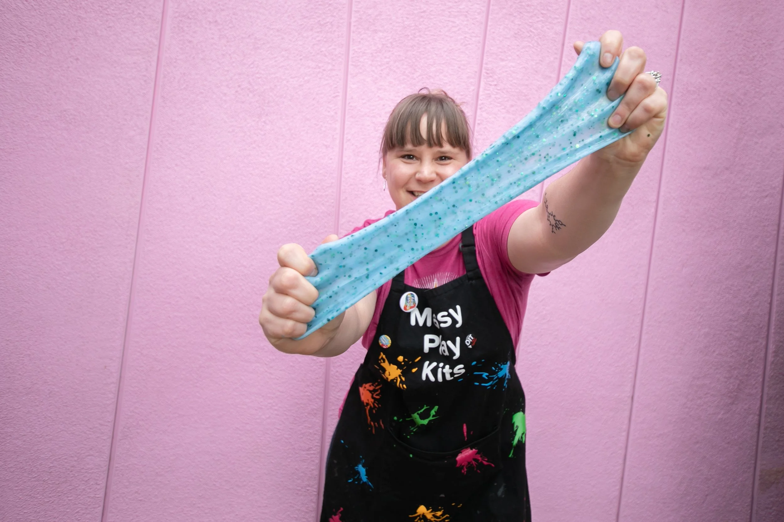 Robin Imholte, the Messy Play Lady, stretching blue glitter slime in front of a pink wall