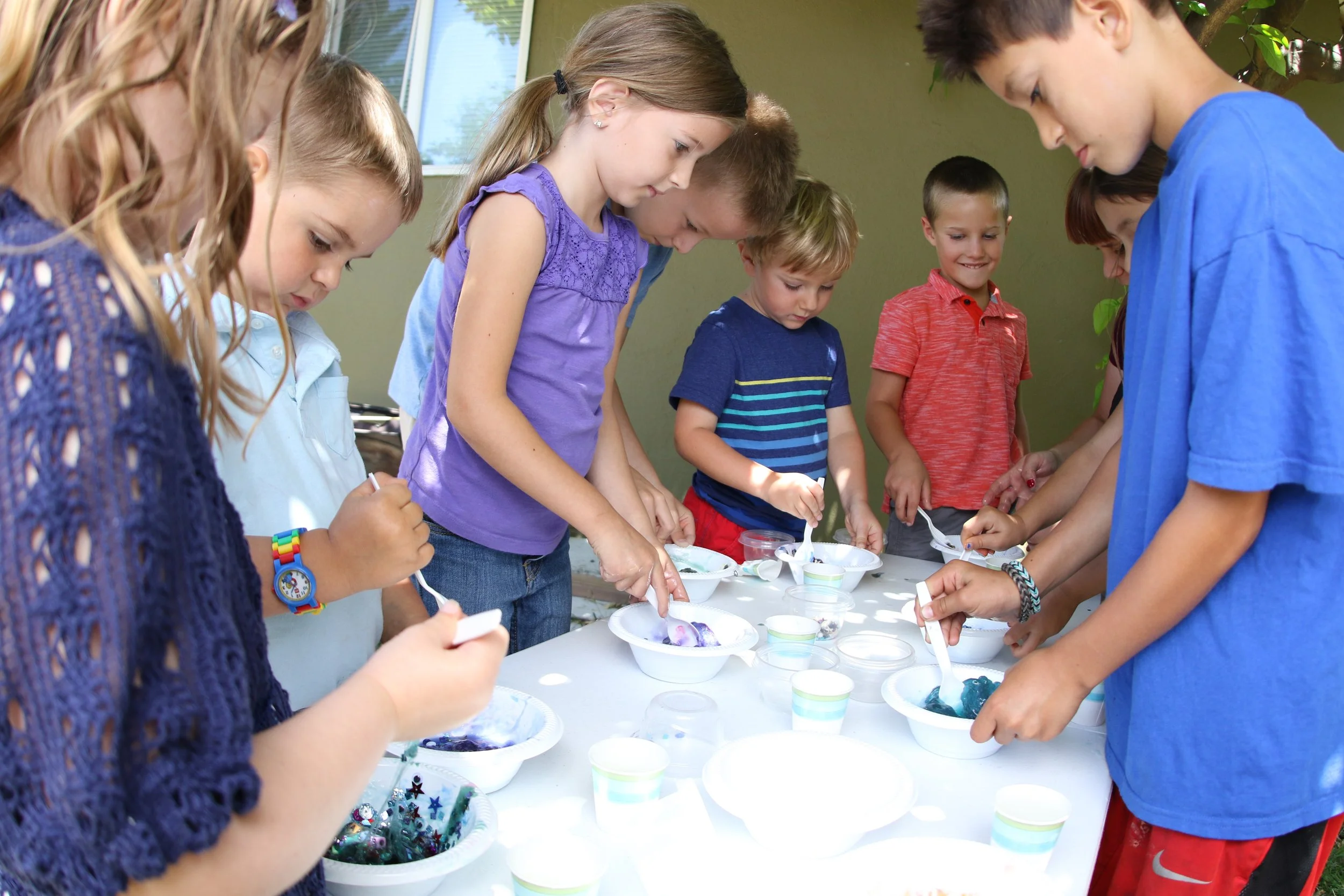 Children and adults enjoy a colorful outdoor art activity with bowls, spoons, and craft materials at a table during a Messy Play Slime Party.