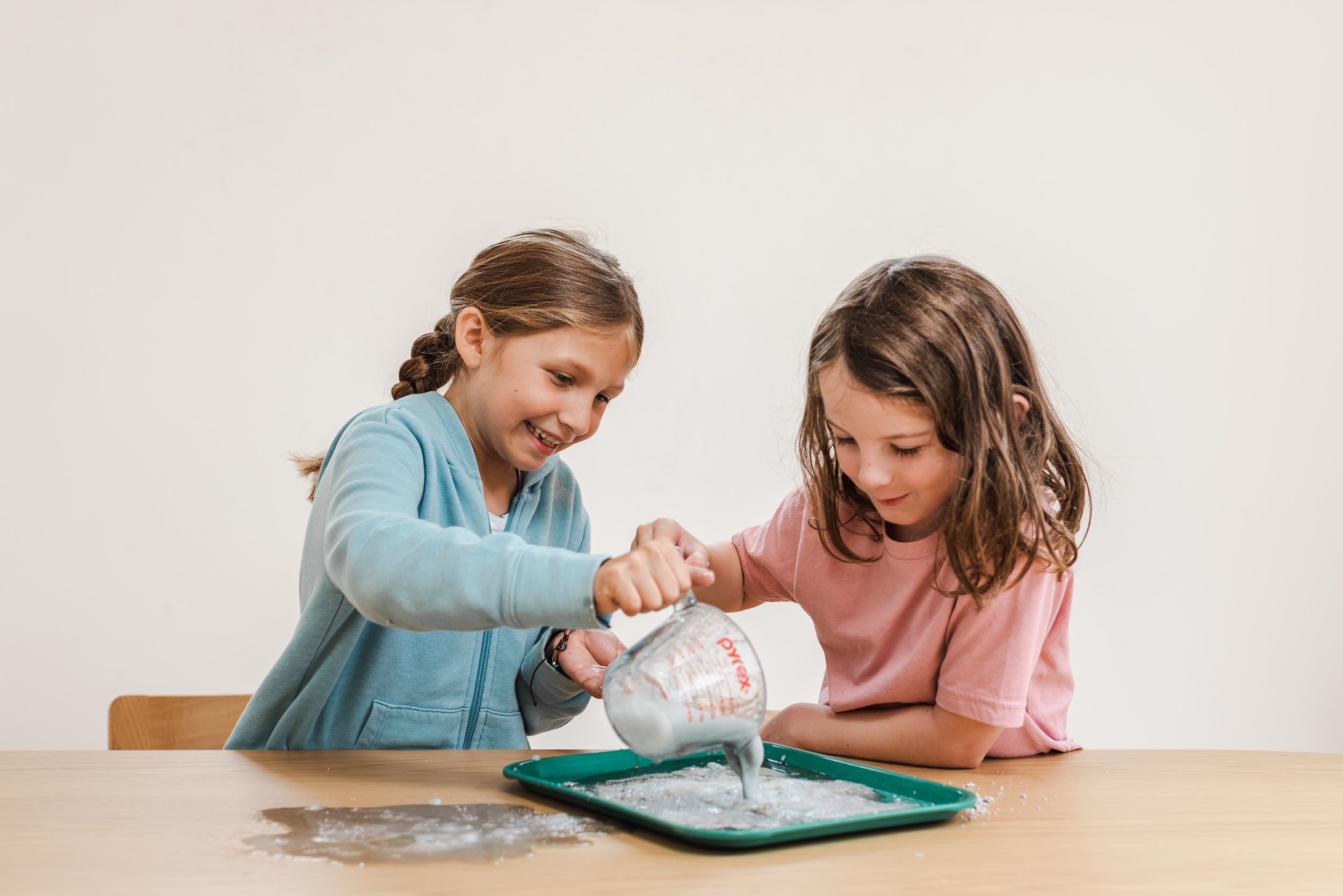 Two girls pour a foamy mixture into a green tray during a hands-on science experiment at a Messy Play Party.