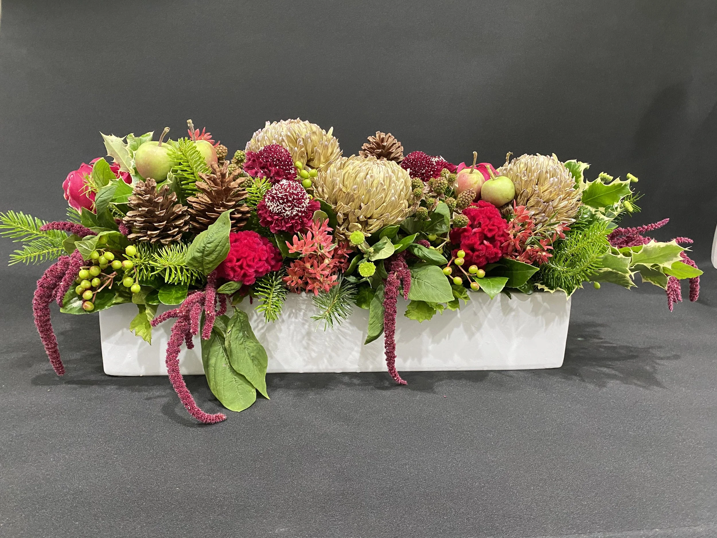 A festive holiday flower arrangement in a white rectangular container, featuring pink and cream flowers, pinecones, green foliage, and small green apples, set against a black background.