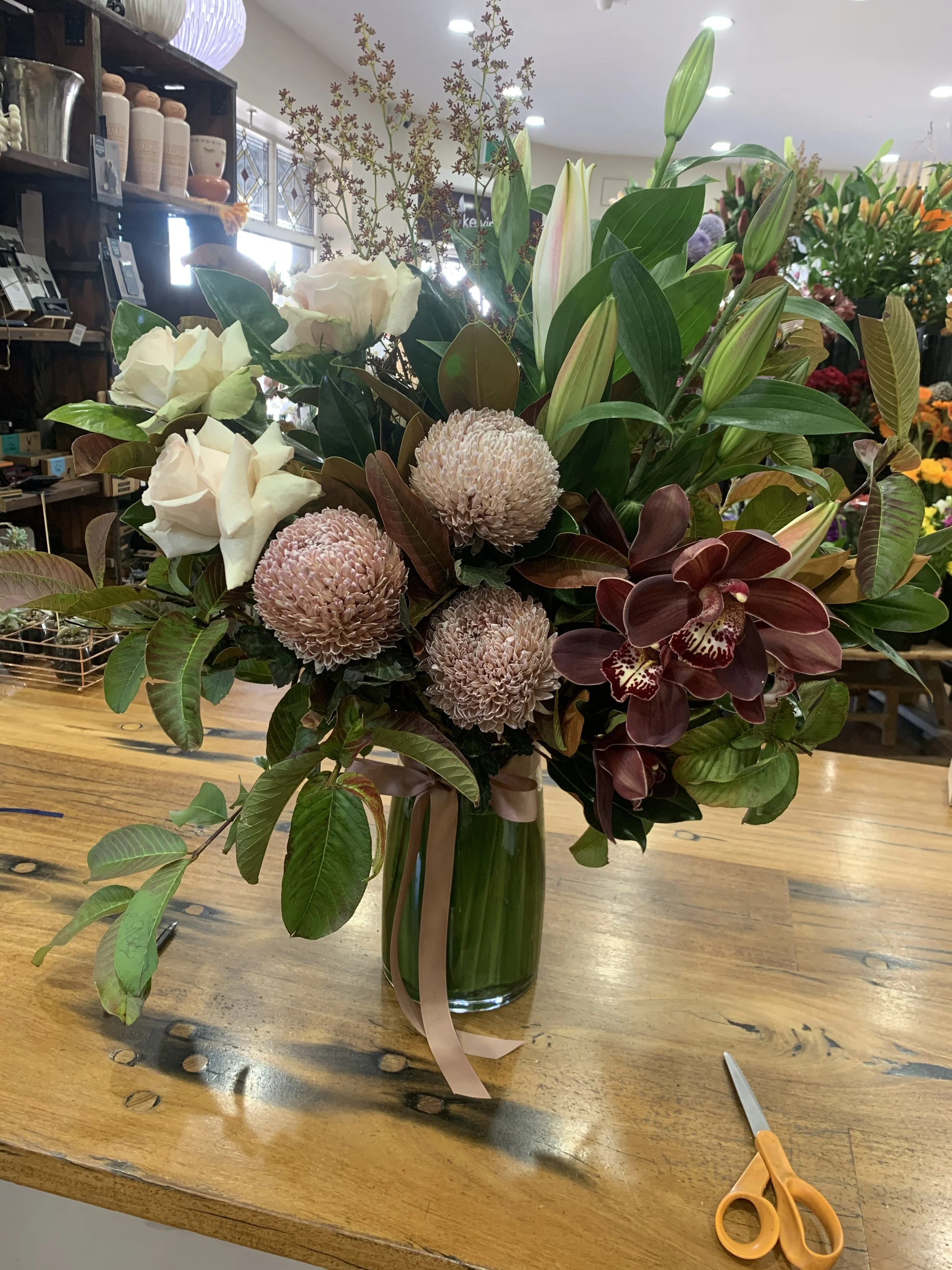 A floral arrangement in a green glass vase on a wooden table, featuring white roses, pink roses, large dusty pink pom-pom flowers, lilies, orchids, and various green leaves, with a pair of orange scissors on the table.