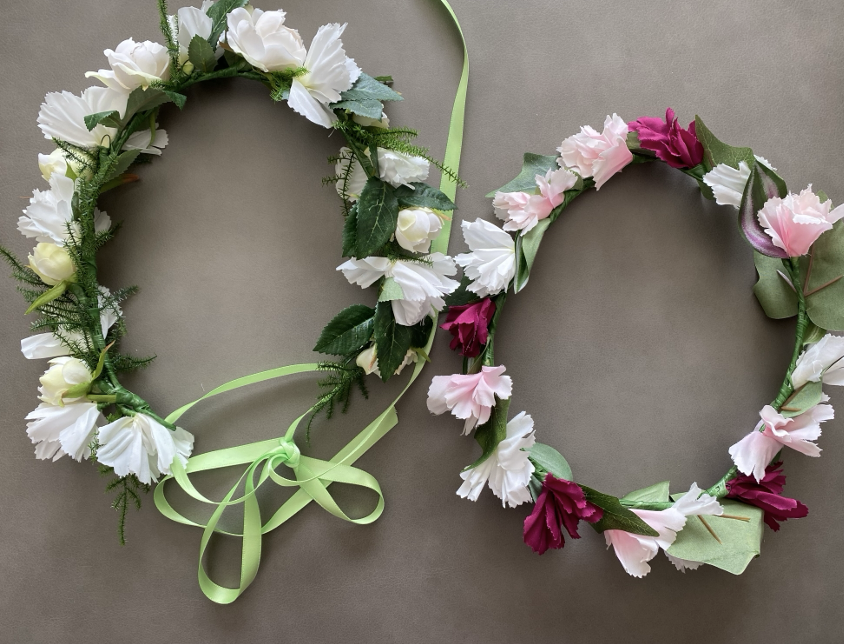 Two floral hair circlets, one with white flowers and green leaves, and the other with pink and burgundy flowers, both decorated with ribbons, on a gray background.