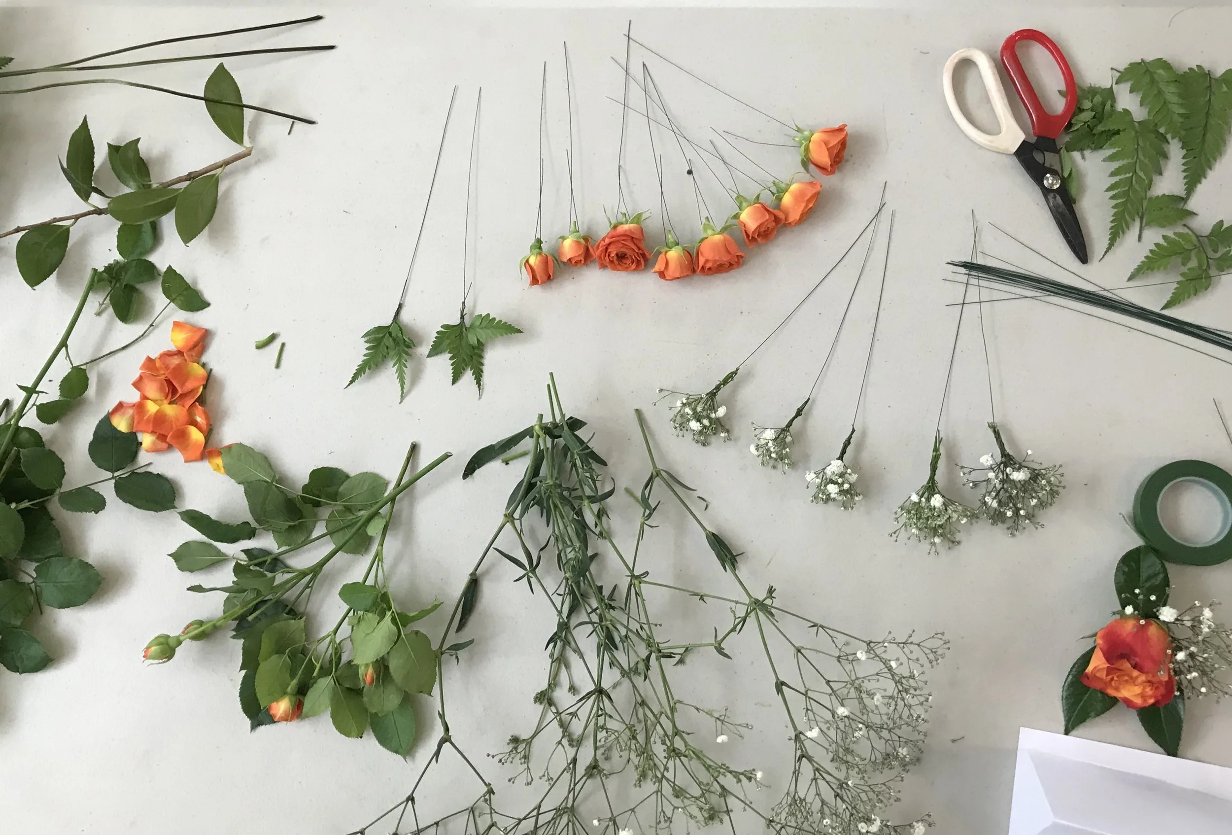 Various flowers, stems, and foliage arranged on a work surface with scissors, tape, and wires.