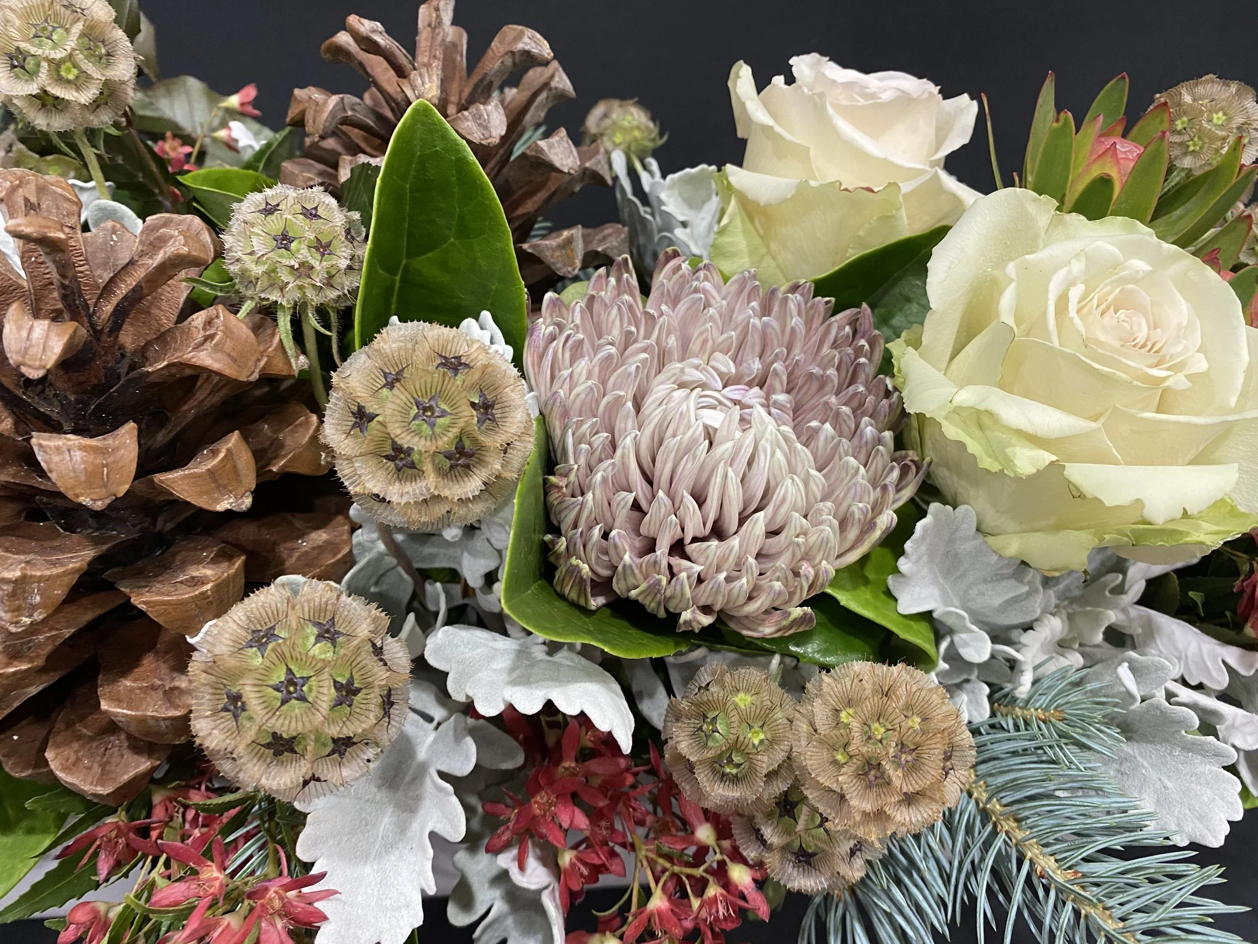 A floral arrangement with white roses, pink and red flowers, pinecones, large green leaves, and various dried and fresh flowers on a black background.