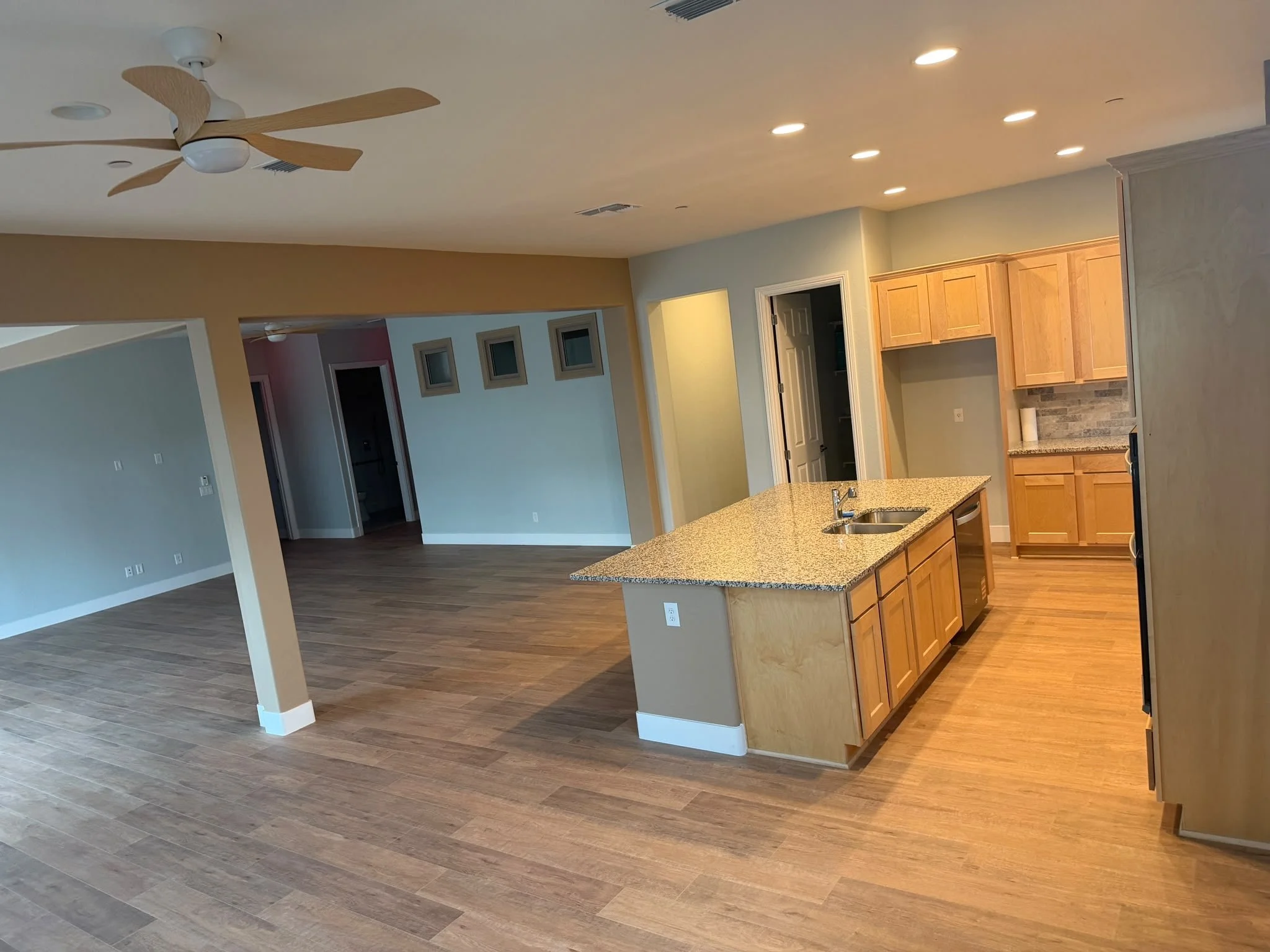 Open-concept living room & kitchen redesign with hardwood floors, light-colored walls, ceiling fan, and recessed lighting. Kitchen with granite countertops and wooden cabinets visible in the background.