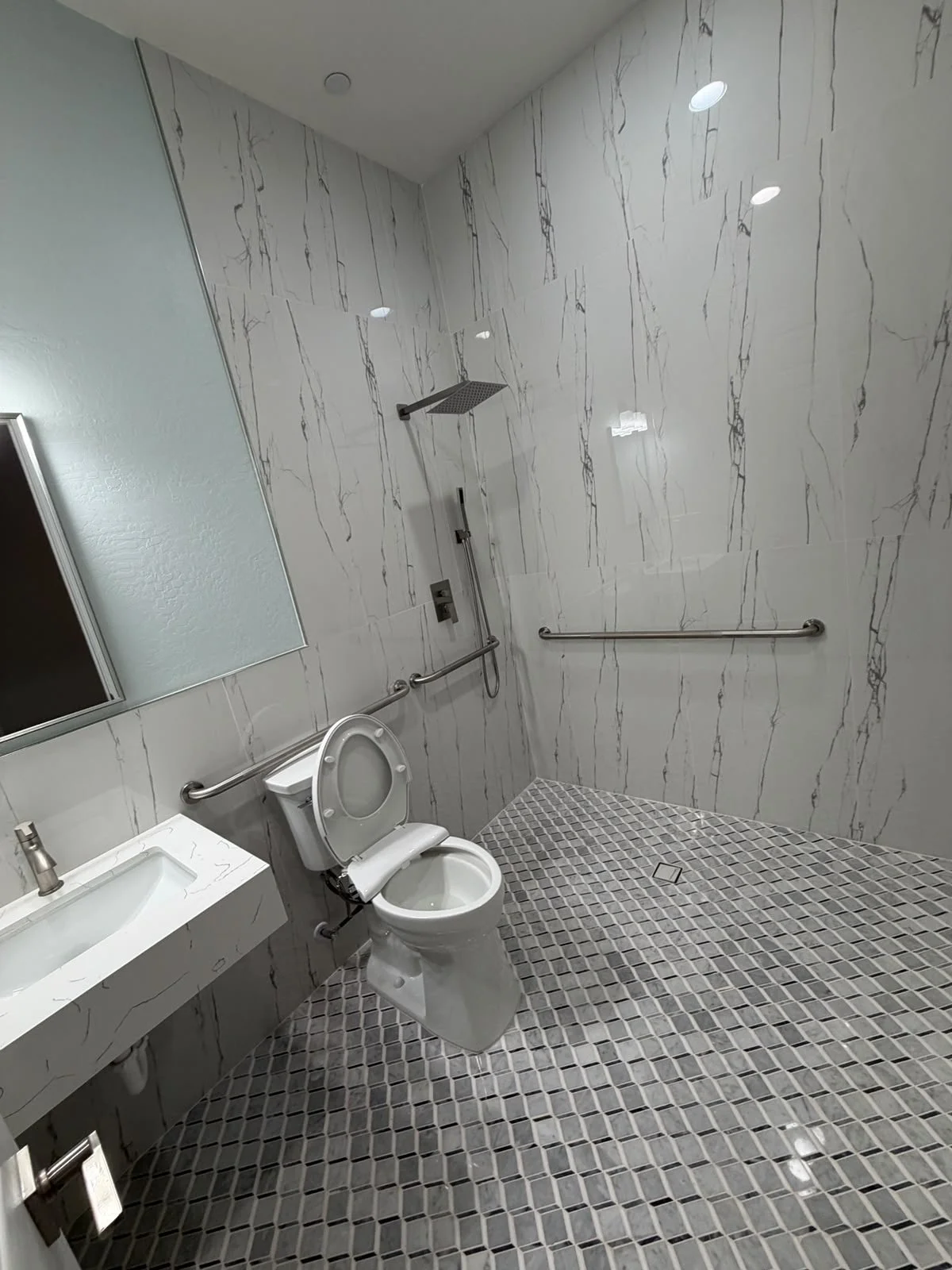 A modern accessible bathroom with marble walls, a walk-in shower with a rain showerhead, a toilet with support bars, a mirror, and a small sink, all with gray and white tile accents.
