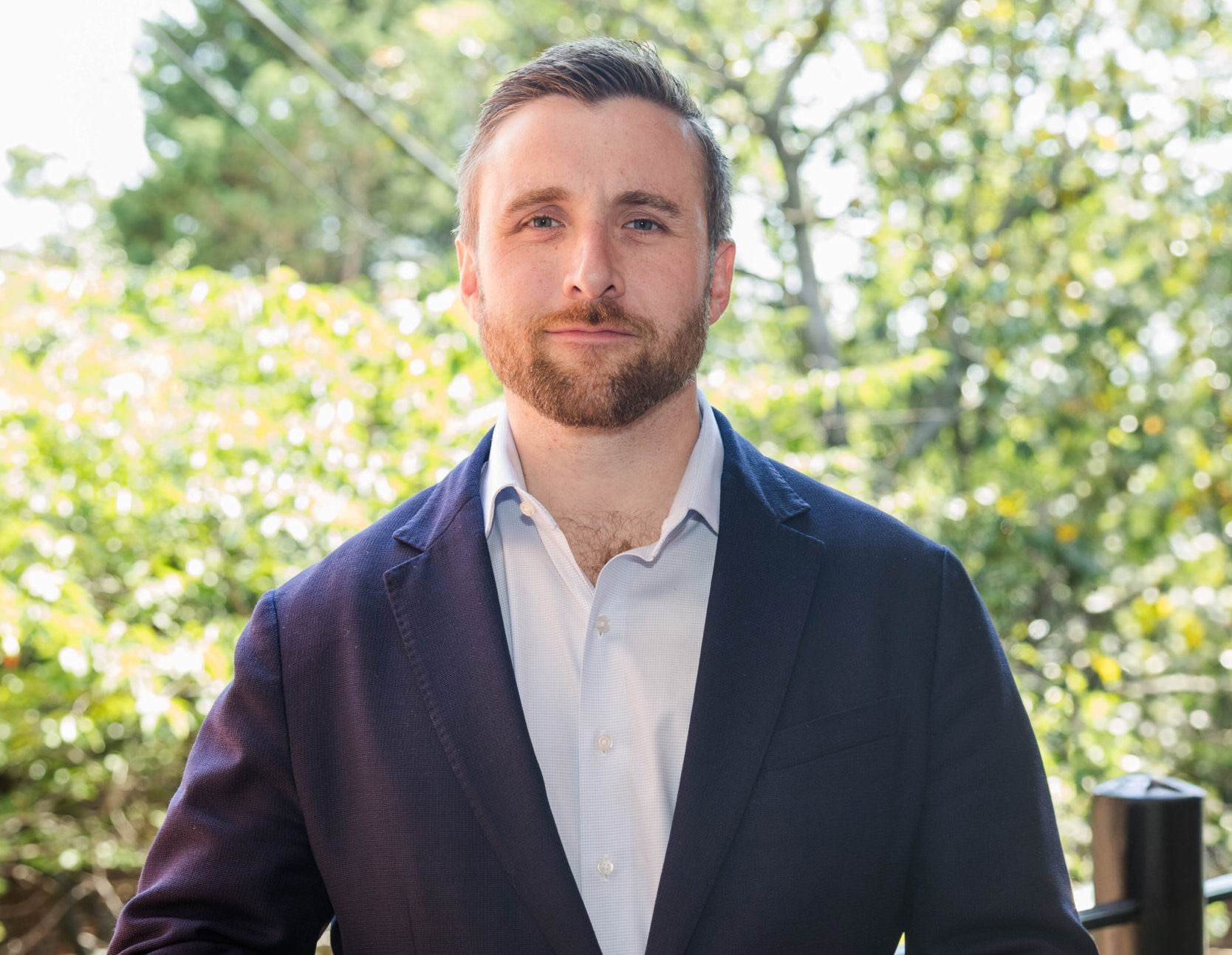 Will Thibeau wearing a white shirt and a dark blue blazer, standing outdoors with green trees and sunlight in the background.