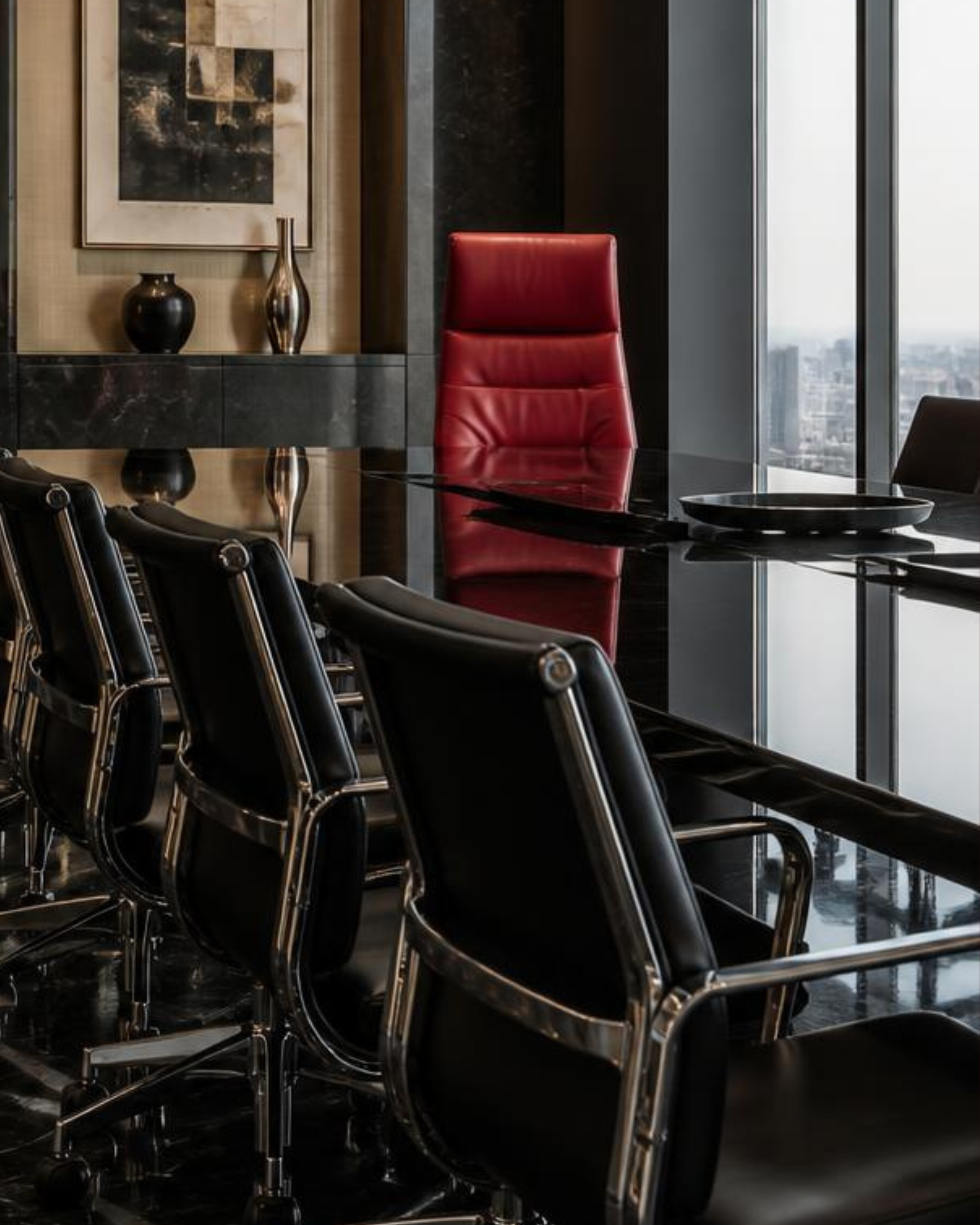 Modern conference room with black table, black chairs, a large red chair, artwork, decorative vases, and large windows showing a cityscape.