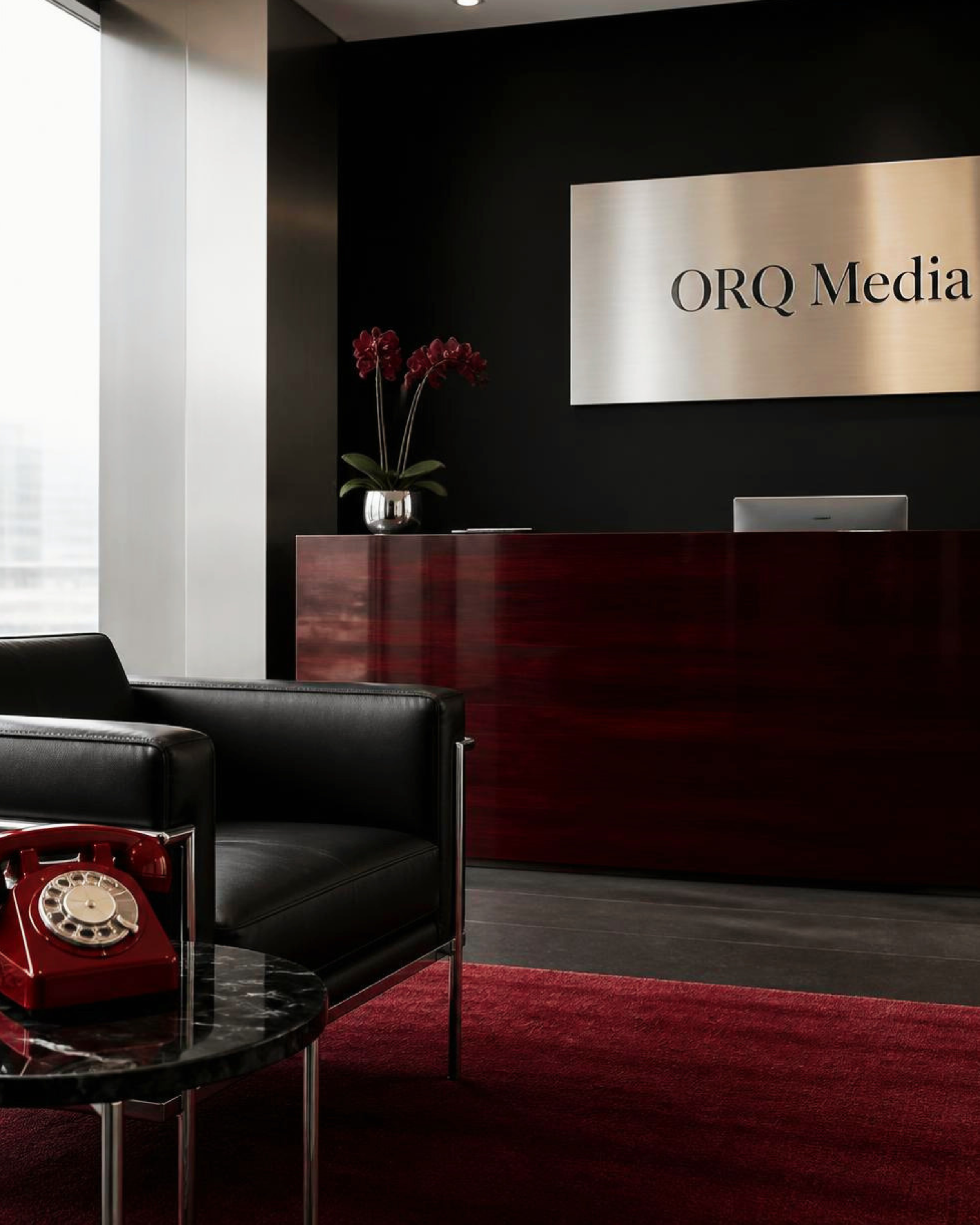Office reception area with a black wall, a silver sign reading 'ORQ Media,' a potted plant with red flowers, a red rotary telephone on a black marble table, and black leather chairs.