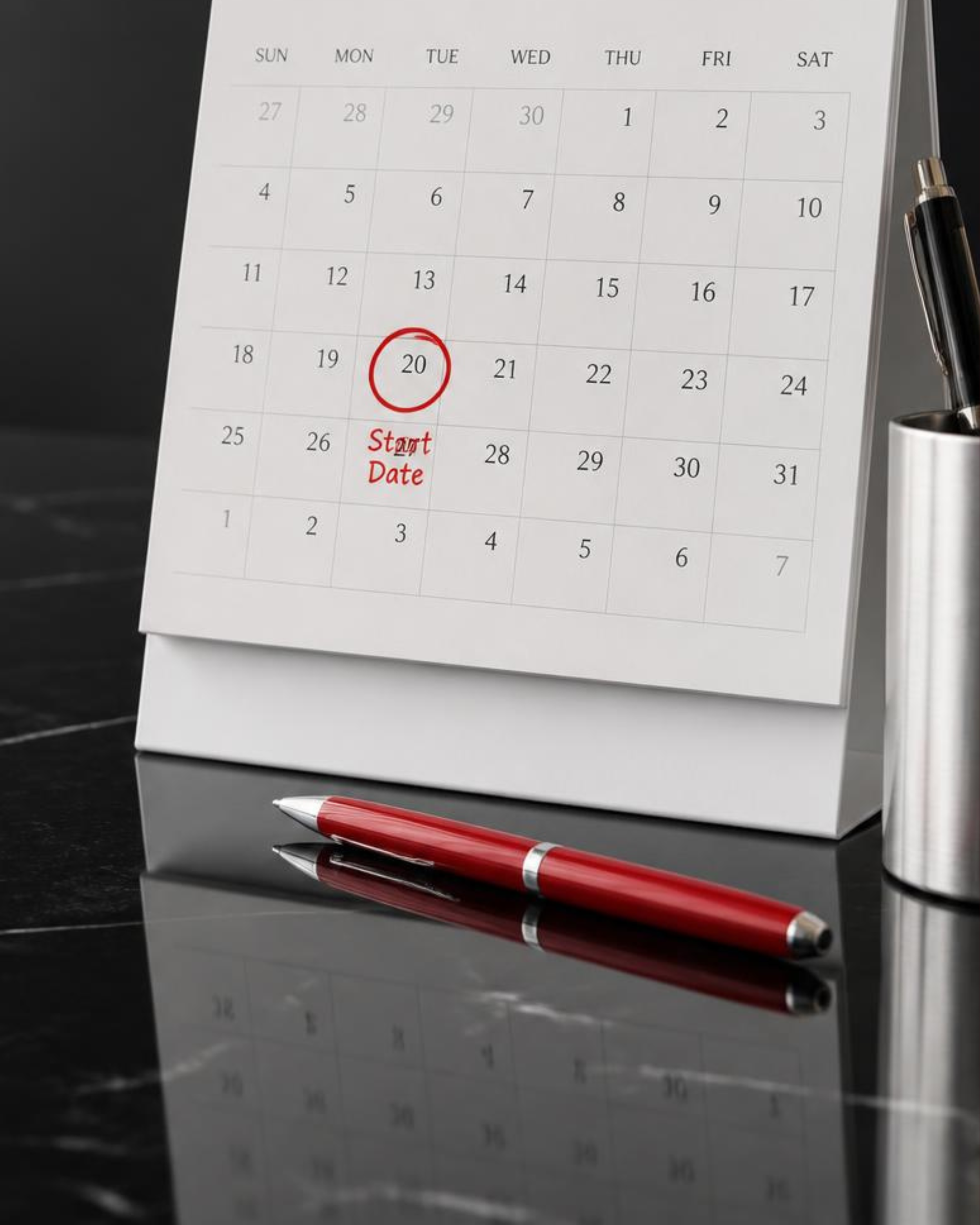 Desk calendar showing the start date of the month on the 20th, with a black pen in a holder and a red pen on a reflective black surface.