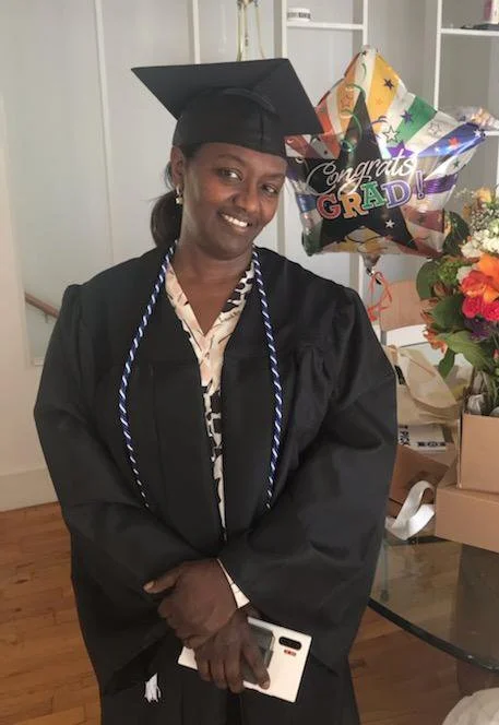 A woman in a graduation cap and gown holding a diploma, smiling, with a colorful graduation balloon and a bouquet of flowers nearby.