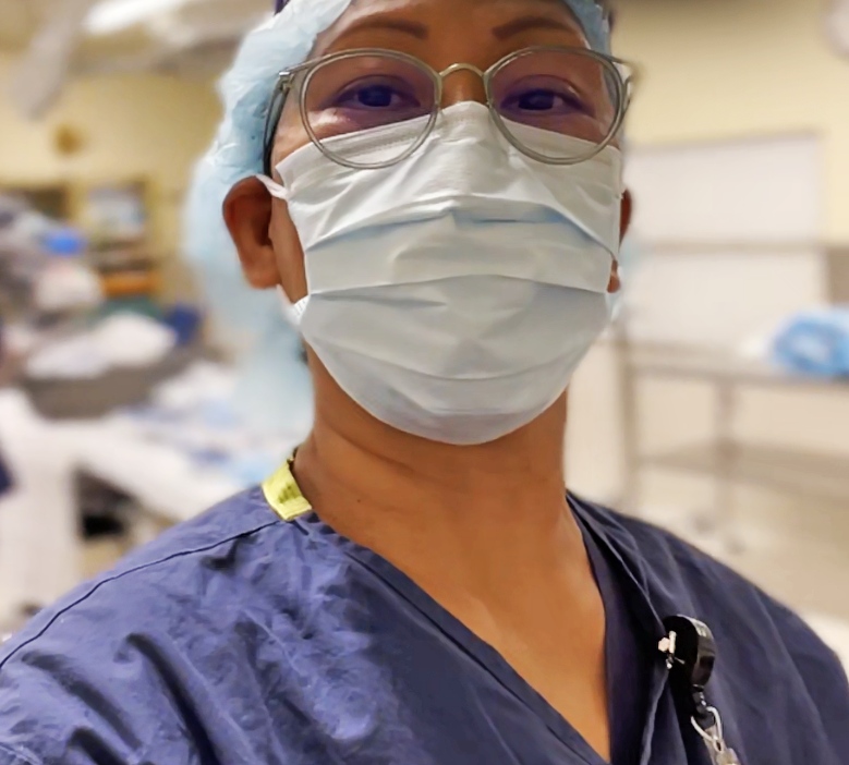 A healthcare worker in scrubs, wearing a surgical mask, glasses, and a hair cap in a hospital setting.