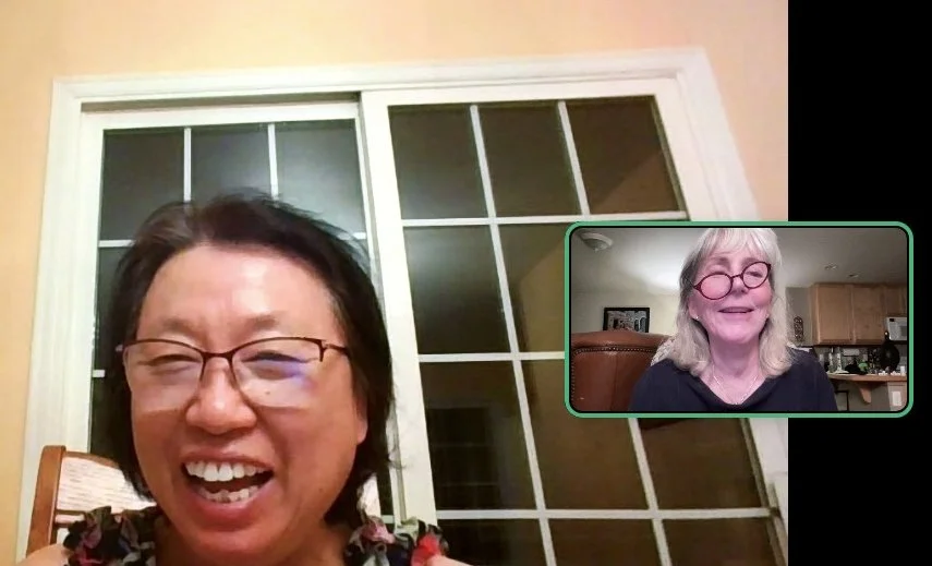 Two women smiling and laughing during a video call. One woman has short black hair and glasses, the other has gray hair and glasses, and is sitting in a kitchen.