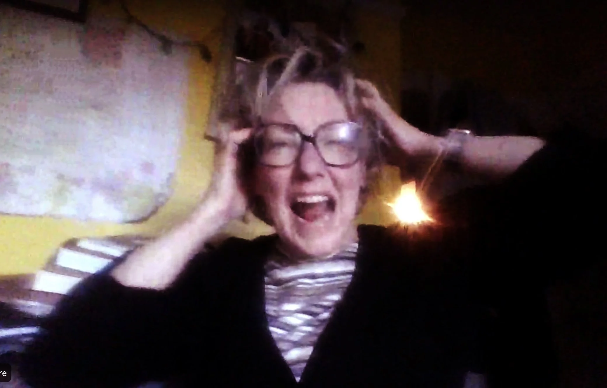 A woman with glasses and striped shirt smiling and holding her hands to her head in a dark room with a glowing light behind her.
