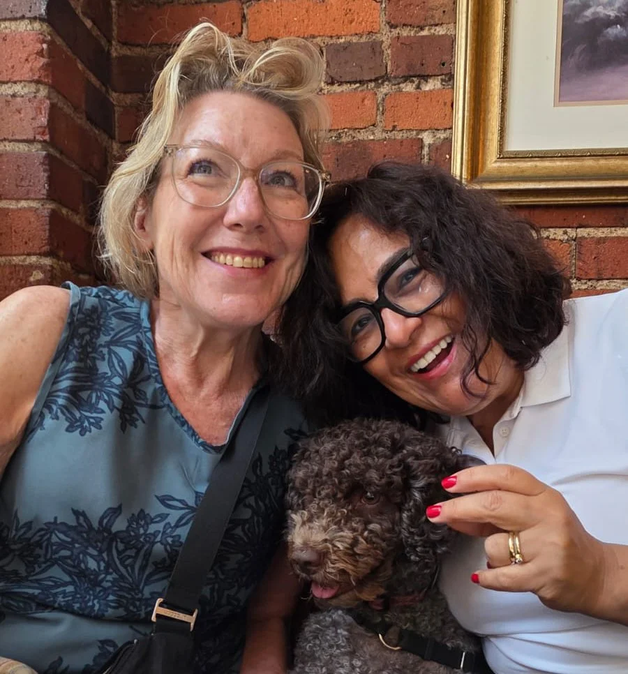 Two women smiling and taking a selfie with a curly-haired dog against a brick wall background.
