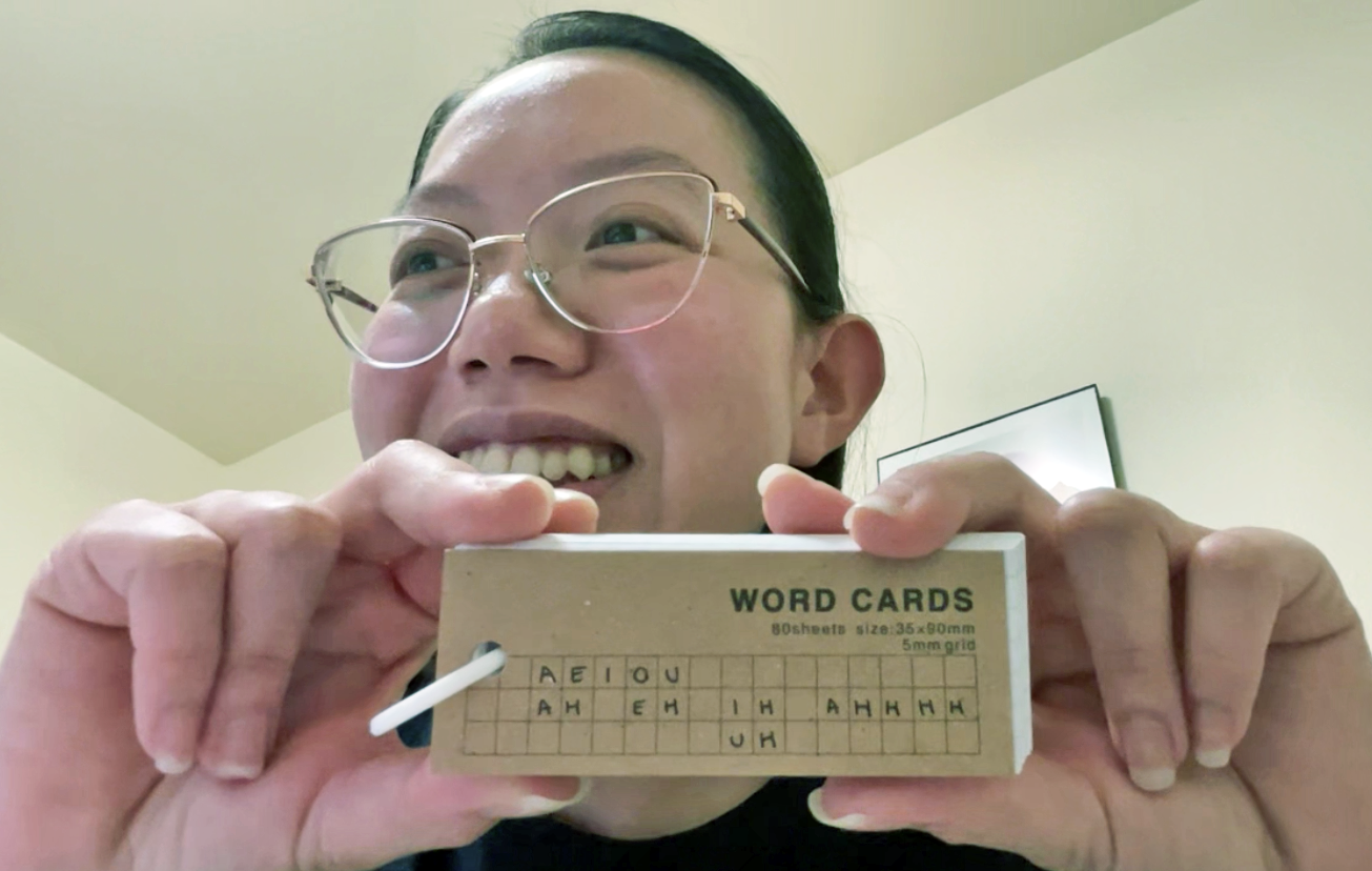 Person with glasses smiling while holding a box of word cards with scrambled letters on a grid.
