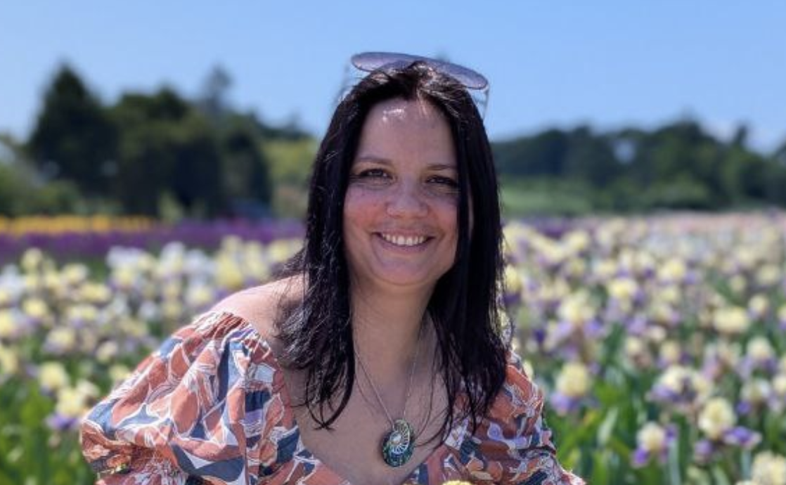 Woman with dark hair, wearing sunglasses on her head and a patterned top, smiling in a flower field on a sunny day.