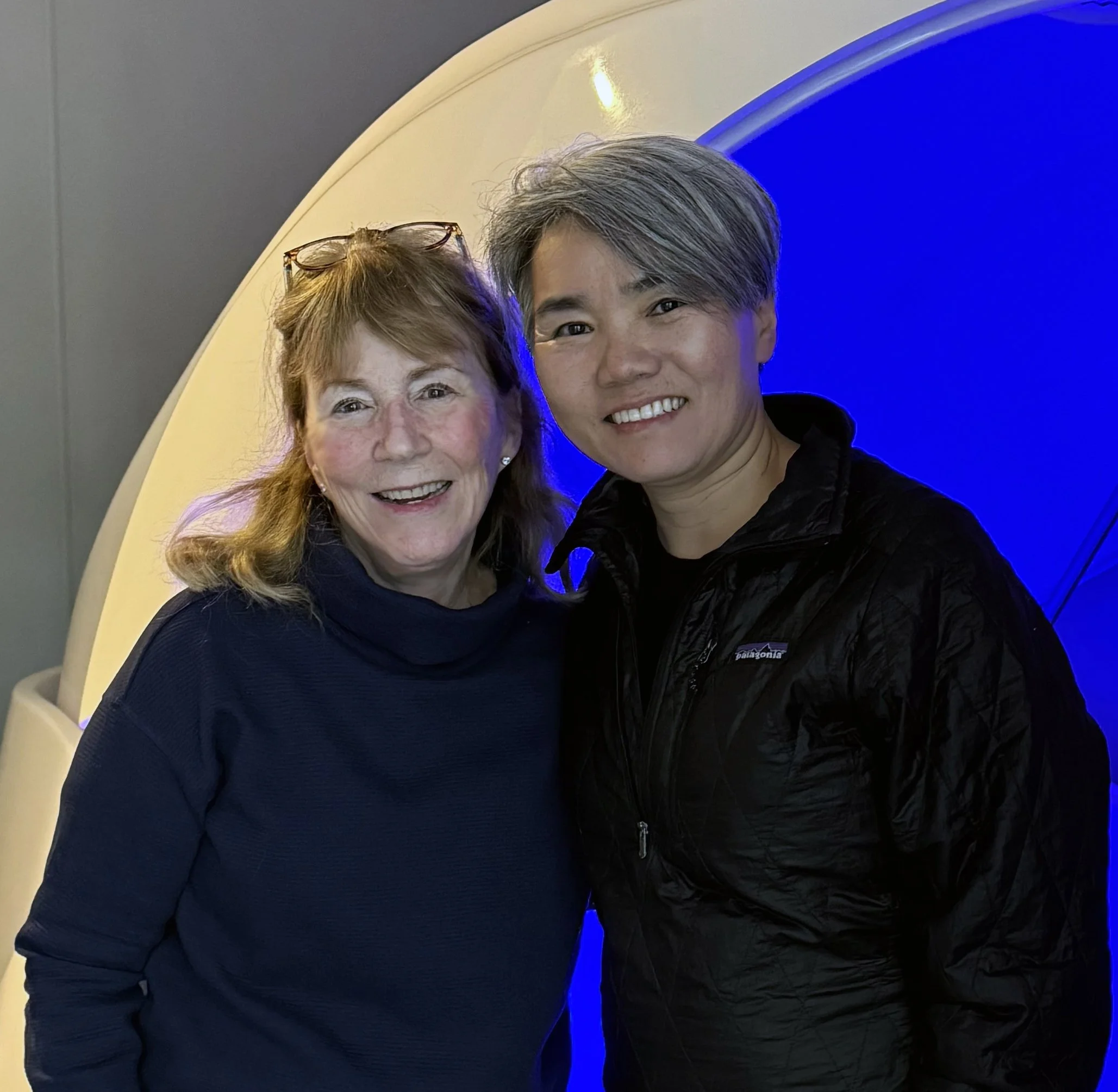 Two people taking a selfie in front of a blue and yellow background, smiling at the camera.
